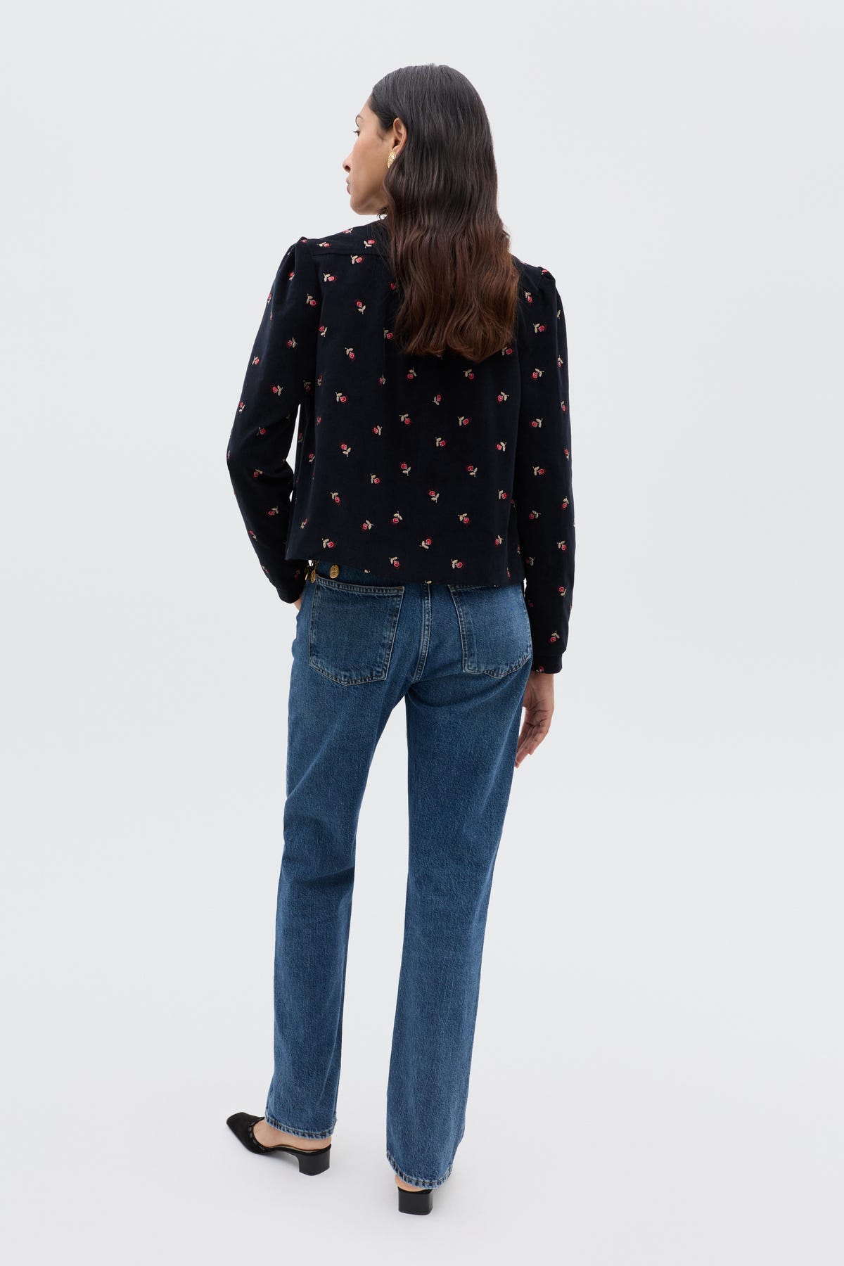 A woman with long brown hair, facing away, wears the Nyla - Dandy Embroidery Deep Navy top with small red floral patterns, cropped blue straight-leg jeans, and black flats against a plain light background.
