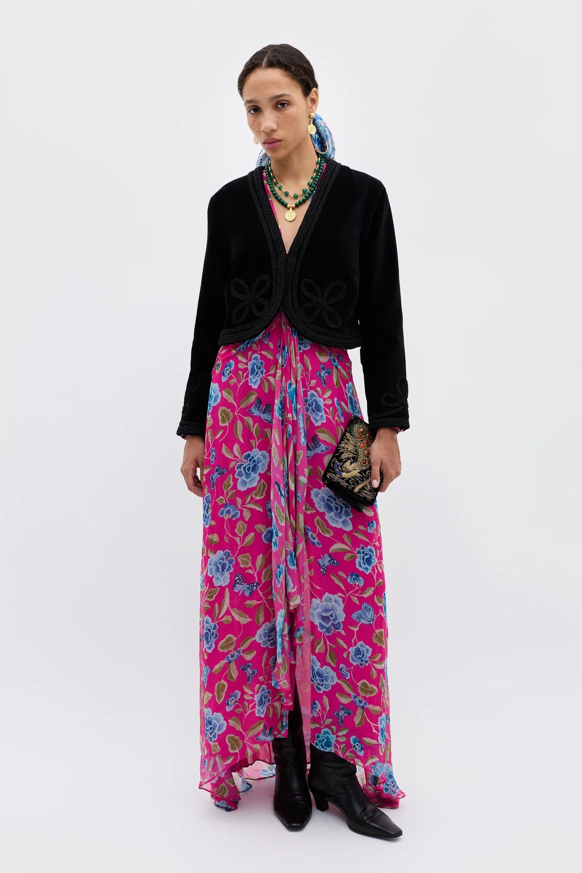 A woman stands against a plain background wearing the Kesia - Clover Appliqué Black jacket over a bright pink floral dress, paired with black boots, layered necklaces, and her hair styled back as she holds a decorative clutch.