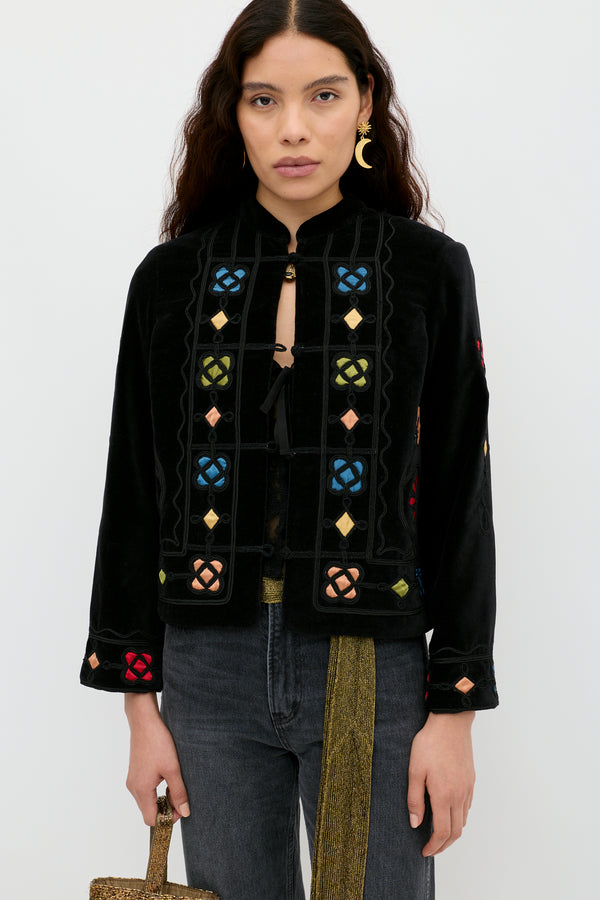 A woman with long wavy hair models the Viktoria - Serino Applique Black jacket, featuring colorful geometric embroidery, over a black top and gray jeans, holding a textured gold handbag against a plain white background.