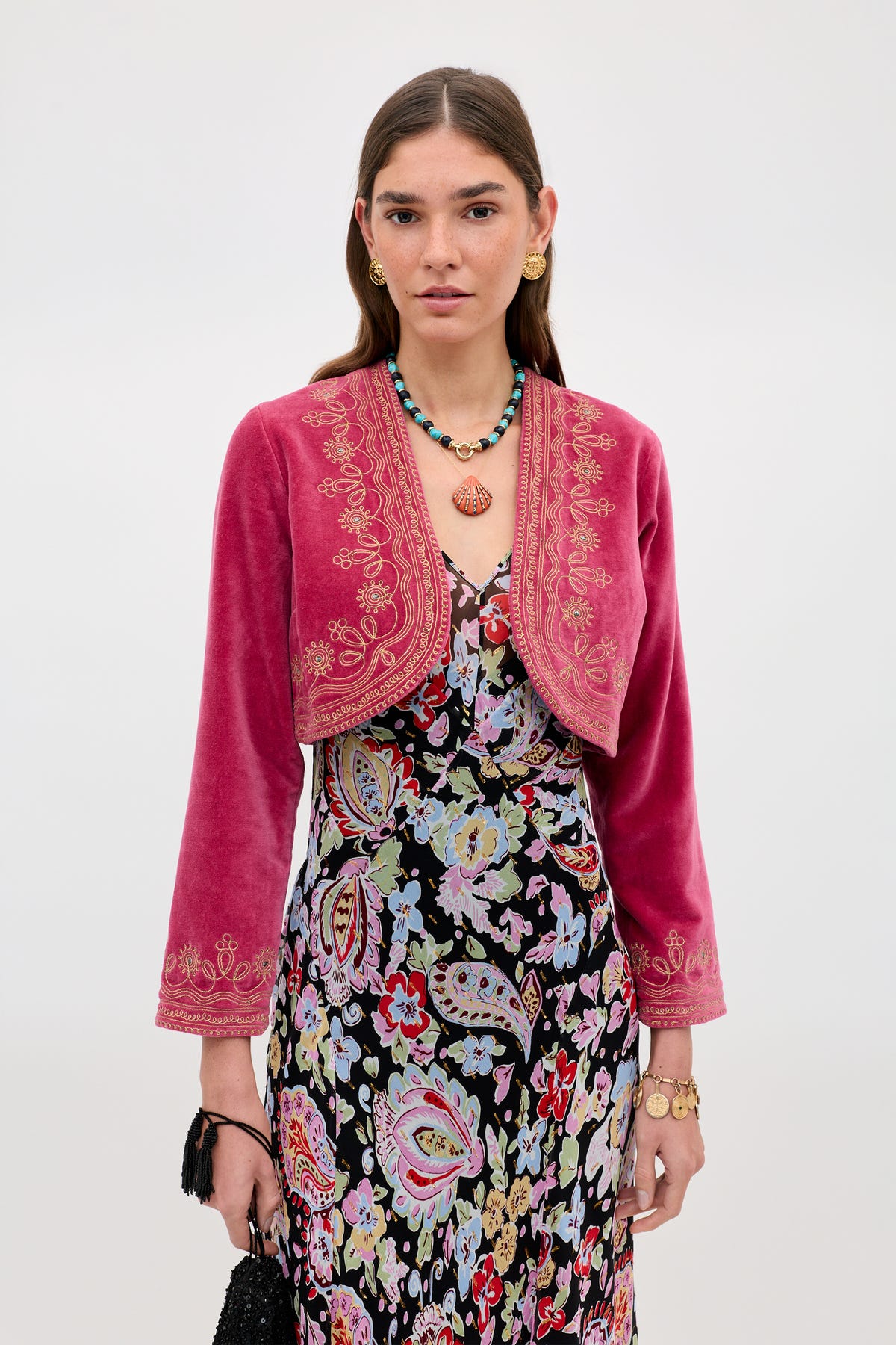 A woman wears the Cliona - Soutache Embroidery Magenta Pink jacket over a floral dress, accessorized with gold jewelry and a black bag, standing against a plain light background.