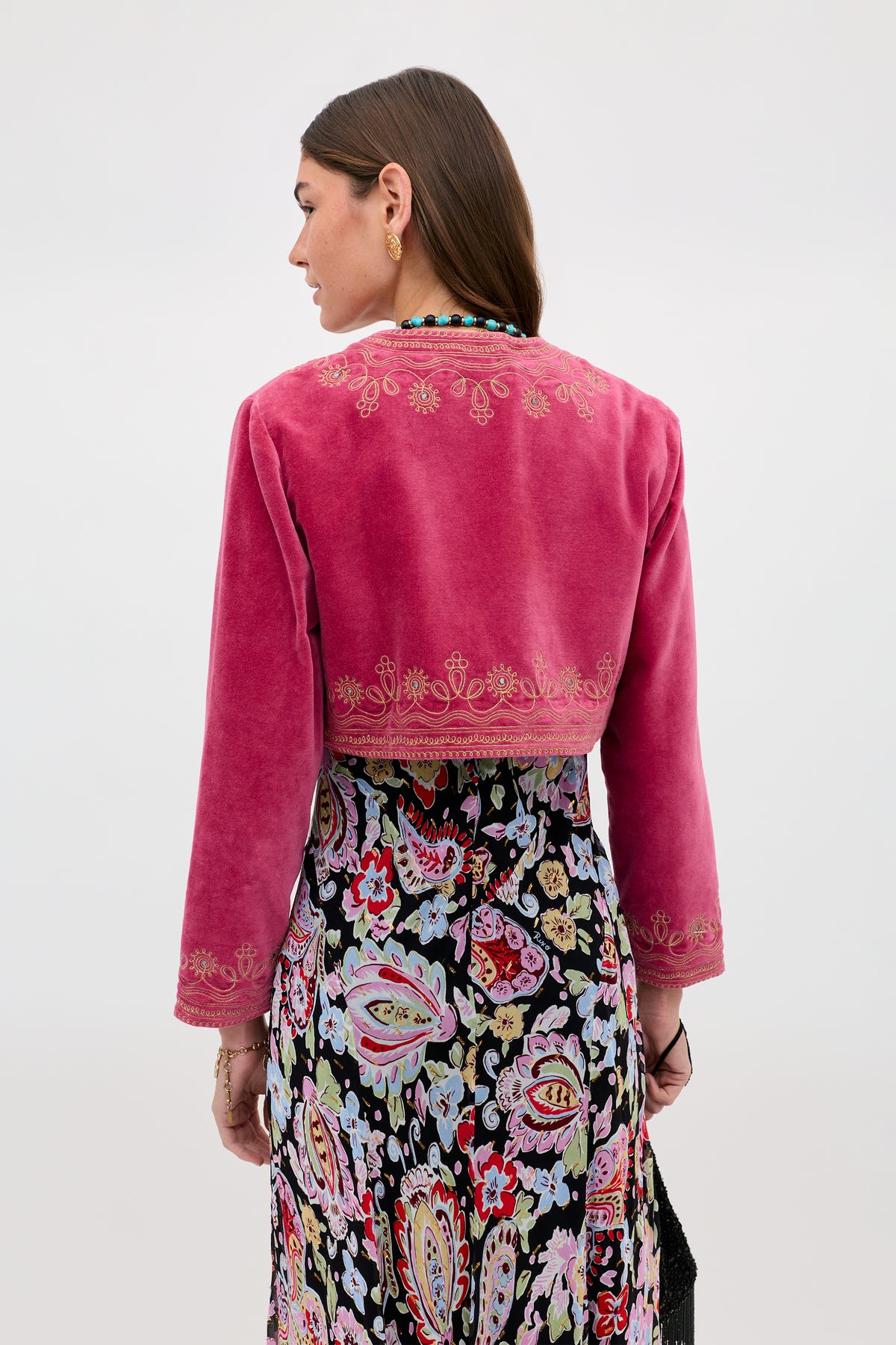 A woman faces away, wearing the Cliona - Soutache Embroidery Magenta Pink velvet jacket with gold detailing and a long, colorful paisley skirt. The backdrop is plain and light-colored.