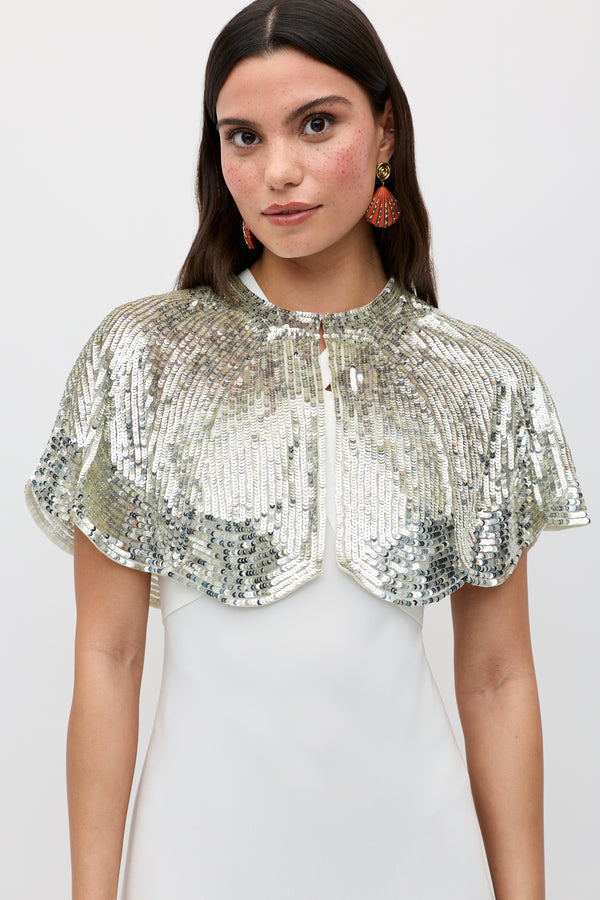 A woman with long dark hair and dangling orange earrings wears the Evienne - Art Deco Embellishment Pearl sequin cape over a white dress, standing against a plain background—an elegant party accessory.