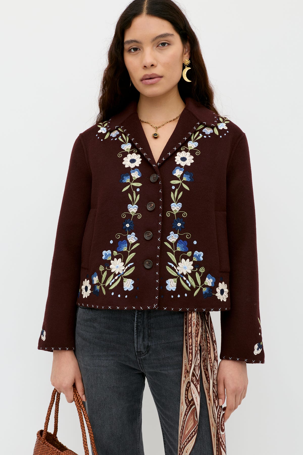A woman with long dark hair wears the Jude - Bliss Embroidery Brown jacket, styled with black jeans, gold jewelry, a brown woven bag, and a patterned scarf against a plain white background.