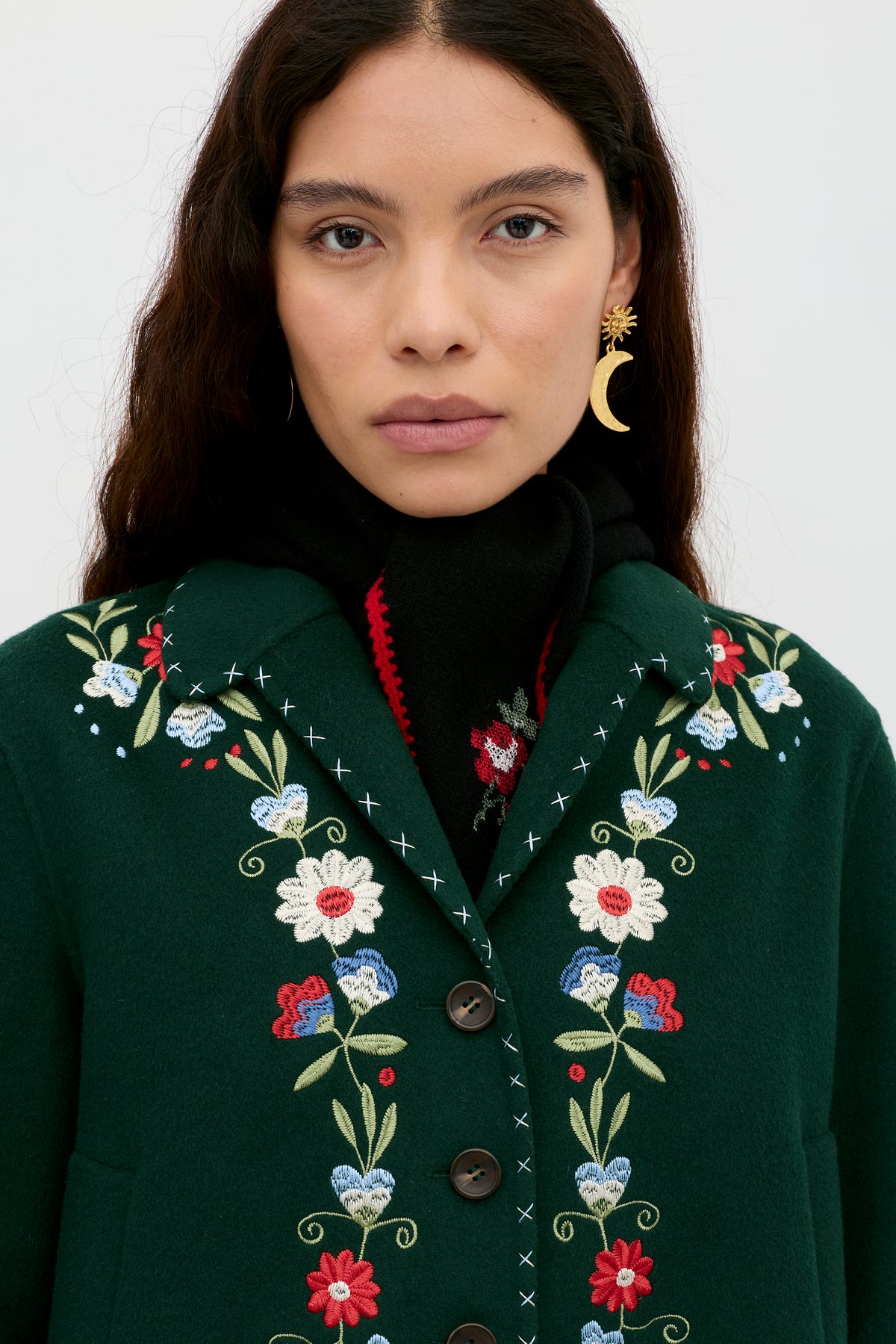 A woman with long brown hair wears the dark green Jude - Bliss Embroidery Green jacket, featuring colorful embroidery, a black scarf, and crescent moon-shaped earrings, looking directly at the camera against a plain background.