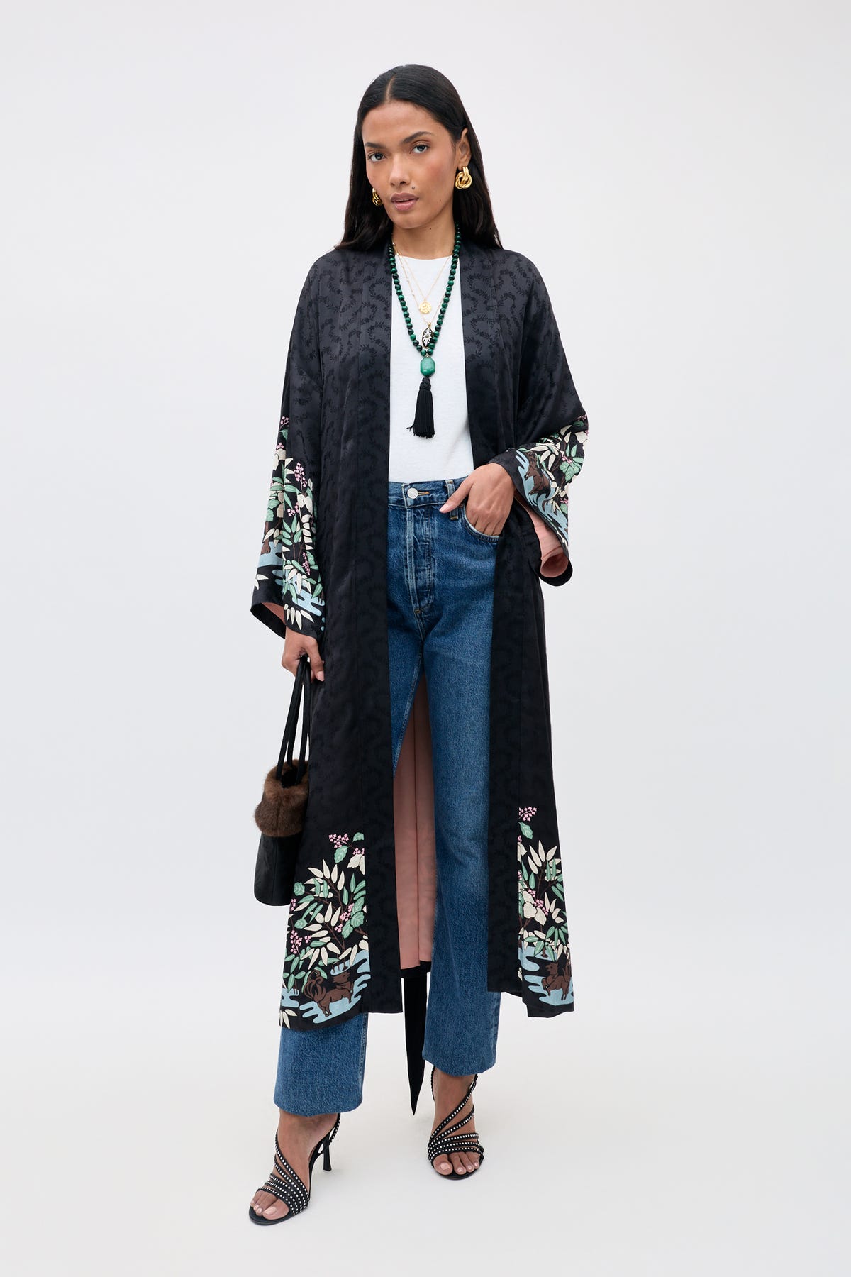Wearing the Kaiden - Blossom Jungle Black kimono over a white top and blue jeans, she completes her look with strappy heels, a black handbag, layered necklaces, and straight dark hair.