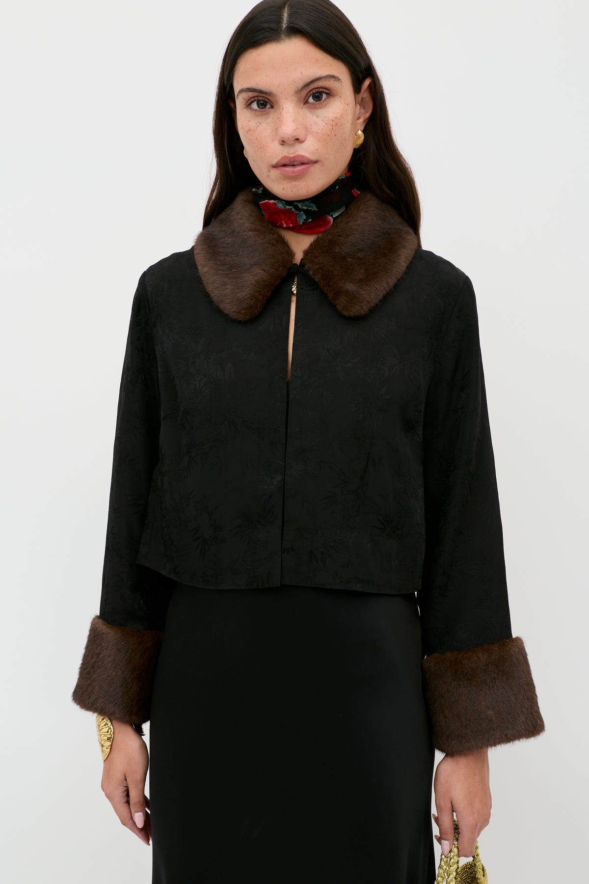 A woman stands against a plain background, wearing the Luanne - Black jacket with a brown fur collar and fur-trimmed cuffs. She pairs it with gold earrings, a floral neck scarf, long dark hair, and holds a small yellow clutch.