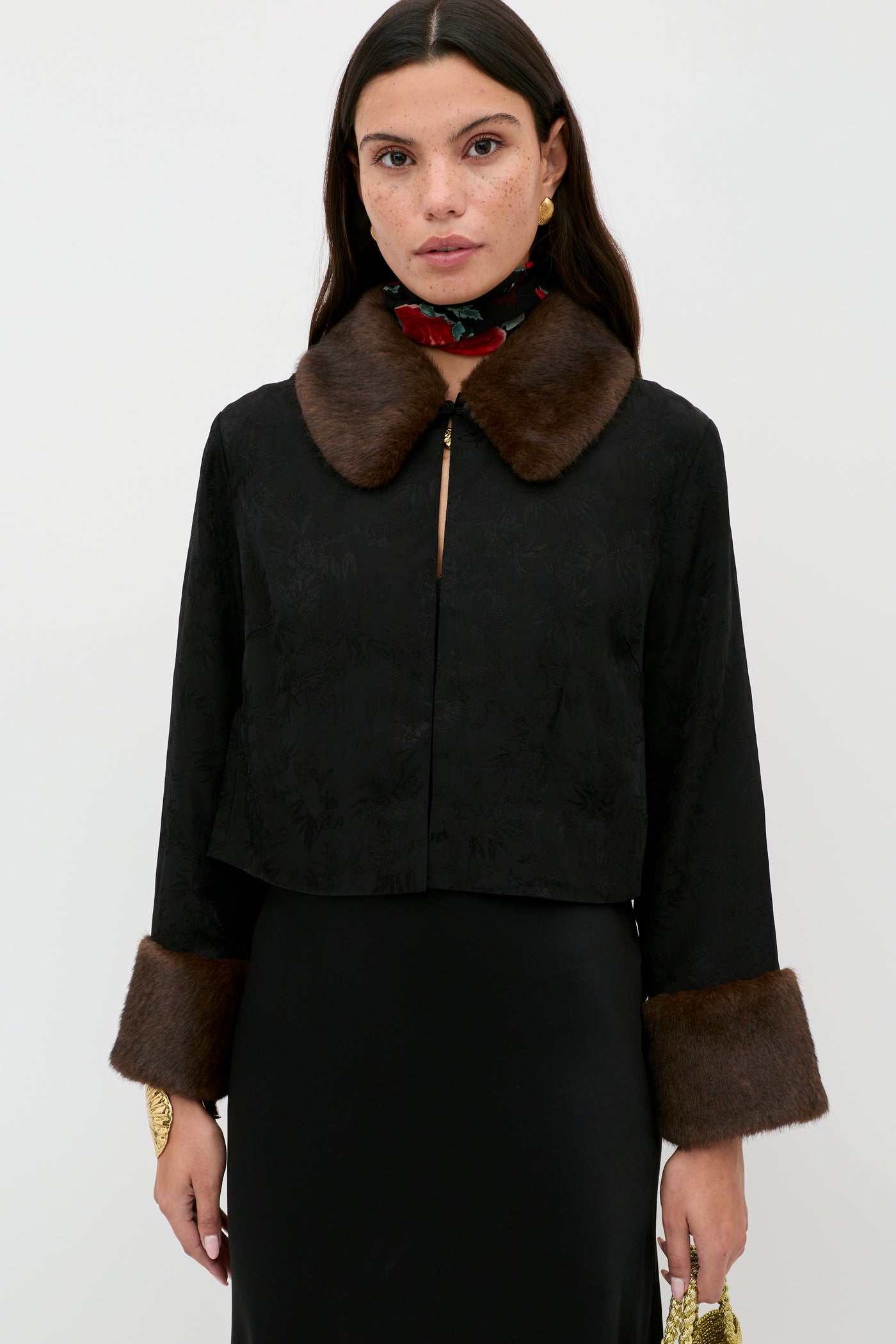 A woman stands against a plain background, wearing the Luanne - Black jacket with a brown fur collar and fur-trimmed cuffs. She pairs it with gold earrings, a floral neck scarf, long dark hair, and holds a small yellow clutch.