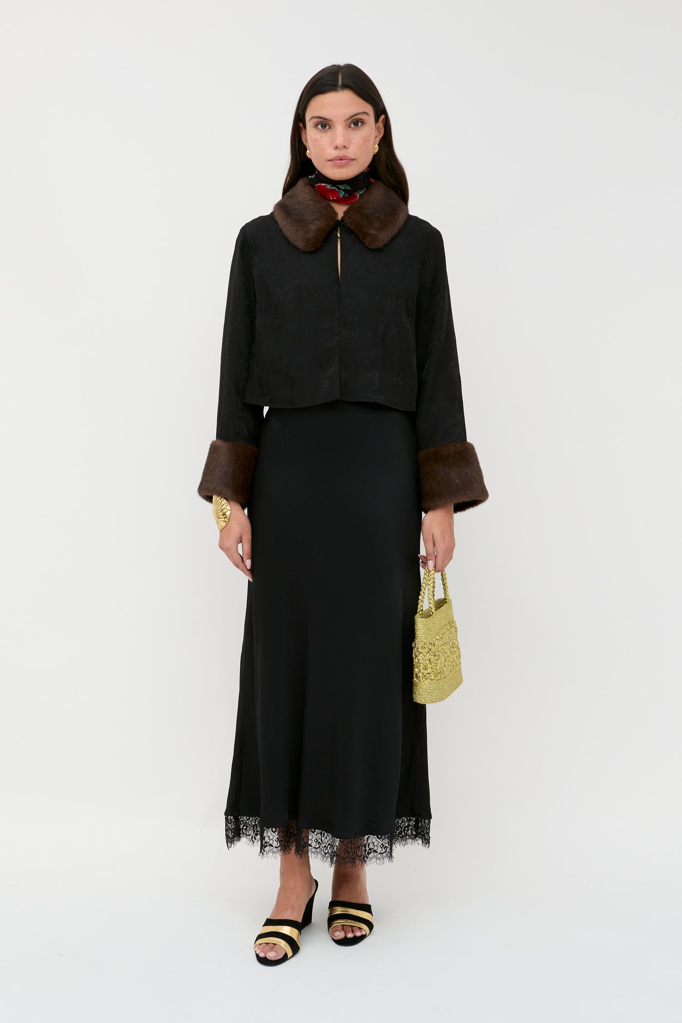 A woman stands against a white background wearing Luanne - Black, a black fur-collared jacket, a long black lace-trim skirt, black and yellow sandals, a red scarf, and holding a yellow textured handbag.