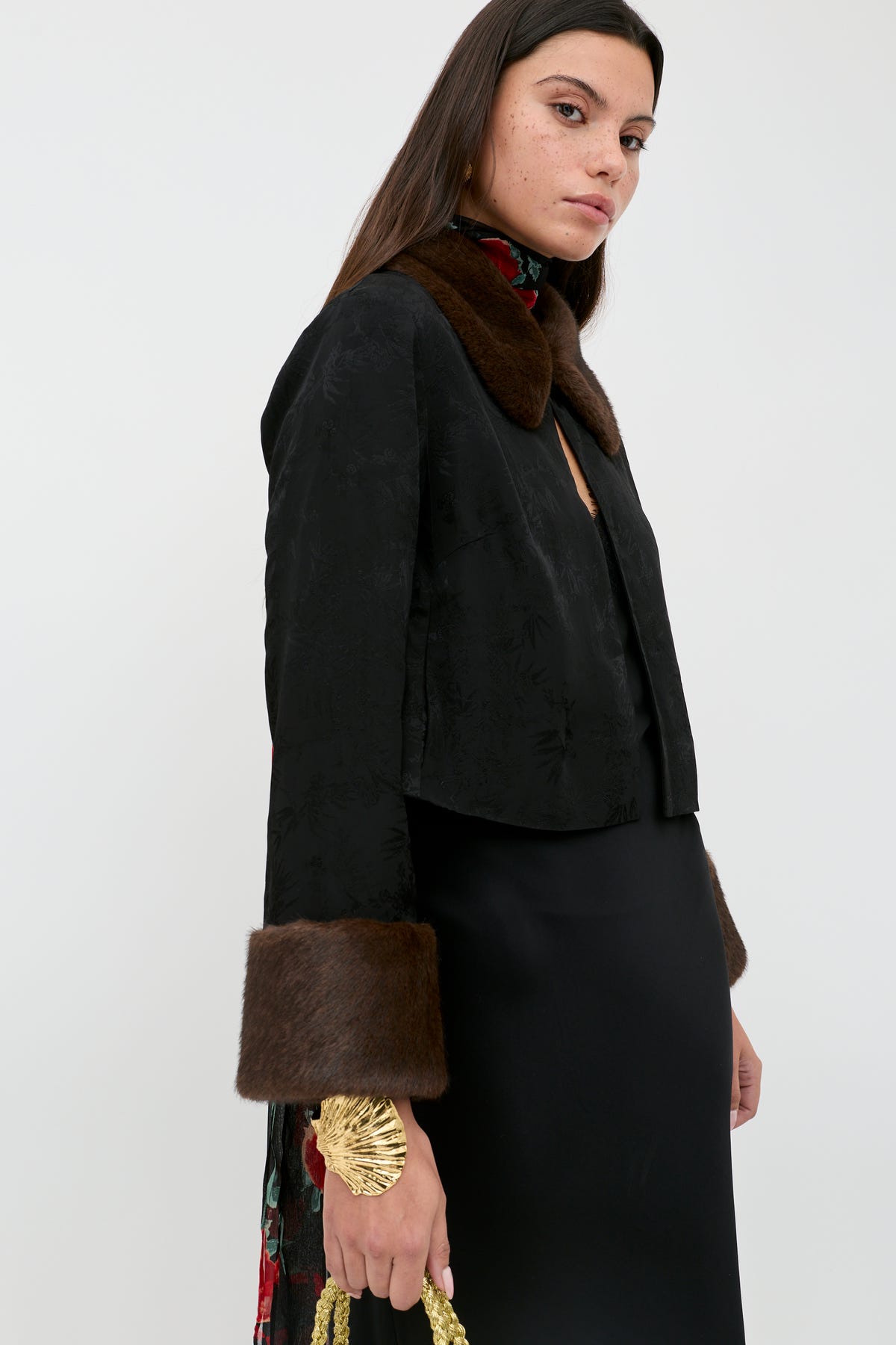 A woman wears the Luanne - Black jacket featuring a brown fur collar and cuffs, paired with a black dress and holding a gold accessory, standing against a plain white background.