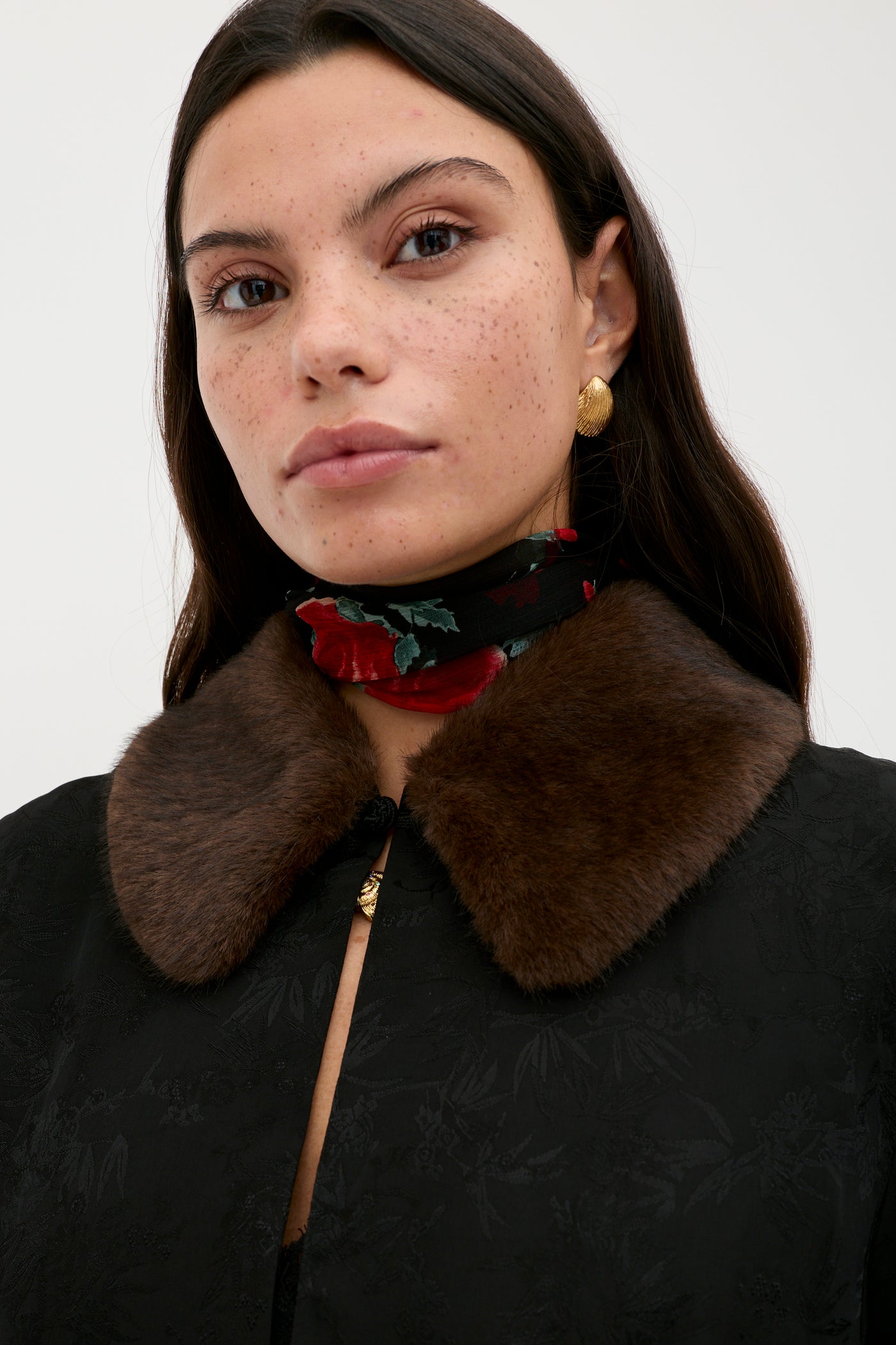 A woman with long dark hair wears the Luanne - Black coat, featuring a textured finish and brown faux fur collar, paired with gold earrings and a red rose floral scarf. She faces the camera with a neutral expression against a plain background.