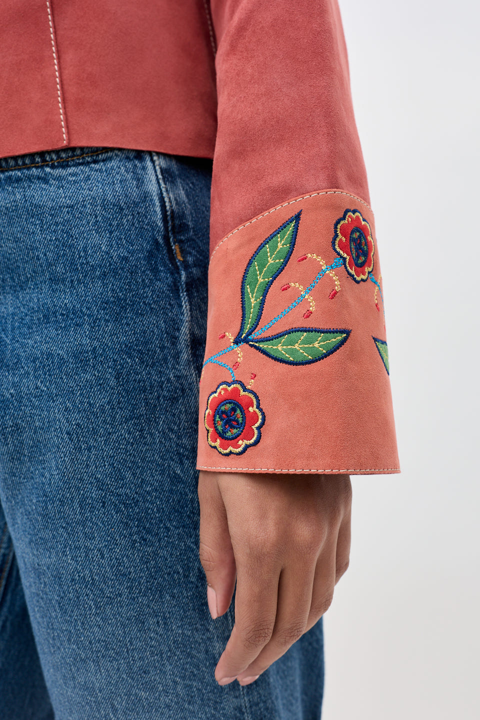A close-up of a person wearing blue jeans and the Karmen - Folk Bloom Appliqué Brown jacket, which features wide sleeves adorned with colorful embroidered flowers and leaves on the sleeve cuff.