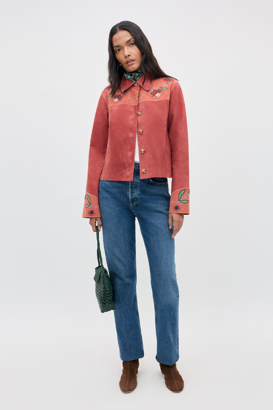 A woman stands against a plain light background wearing a red embroidered jacket, blue jeans, brown boots, and holding the Karmen - Folk Bloom Appliqué Brown handbag.