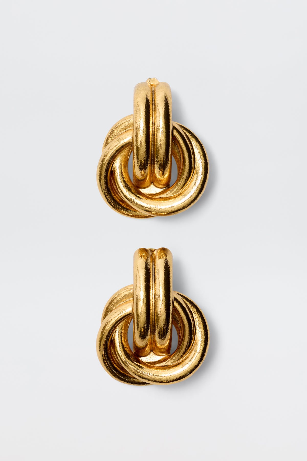 The Nolana - Gold earrings are chunky gold-plated brass hoops with interlocking circular loops, inspired by vintage glamour, shown against a plain light gray background.