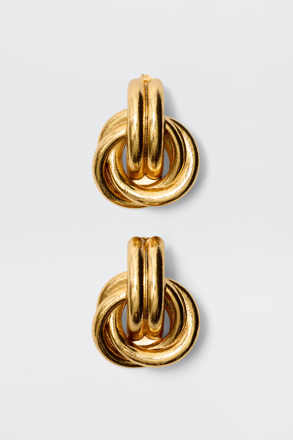 The Nolana - Gold earrings are chunky gold-plated brass hoops with interlocking circular loops, inspired by vintage glamour, shown against a plain light gray background.