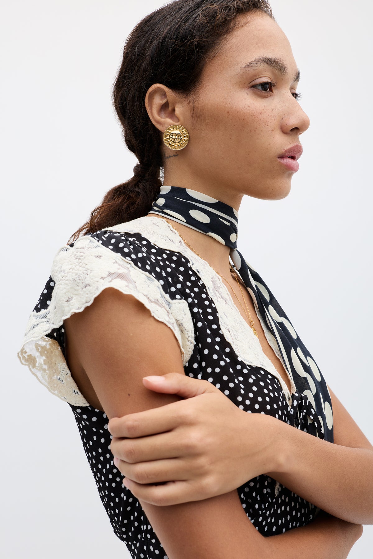 A woman with brown hair in a ponytail wears large round gold-plated brass Noni - Gold earrings, a black and white polka dot top with lace sleeves, and matching scarf, standing against a plain light background.