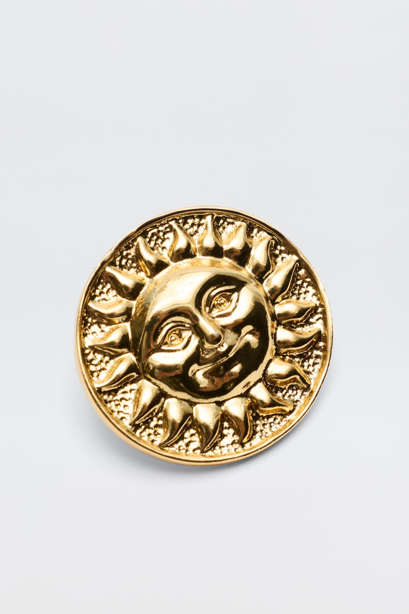 The Noni - Gold is a round gold-plated brass pin with a raised smiling sun face and radiant rays on a textured background—a charming vintage-inspired accessory.