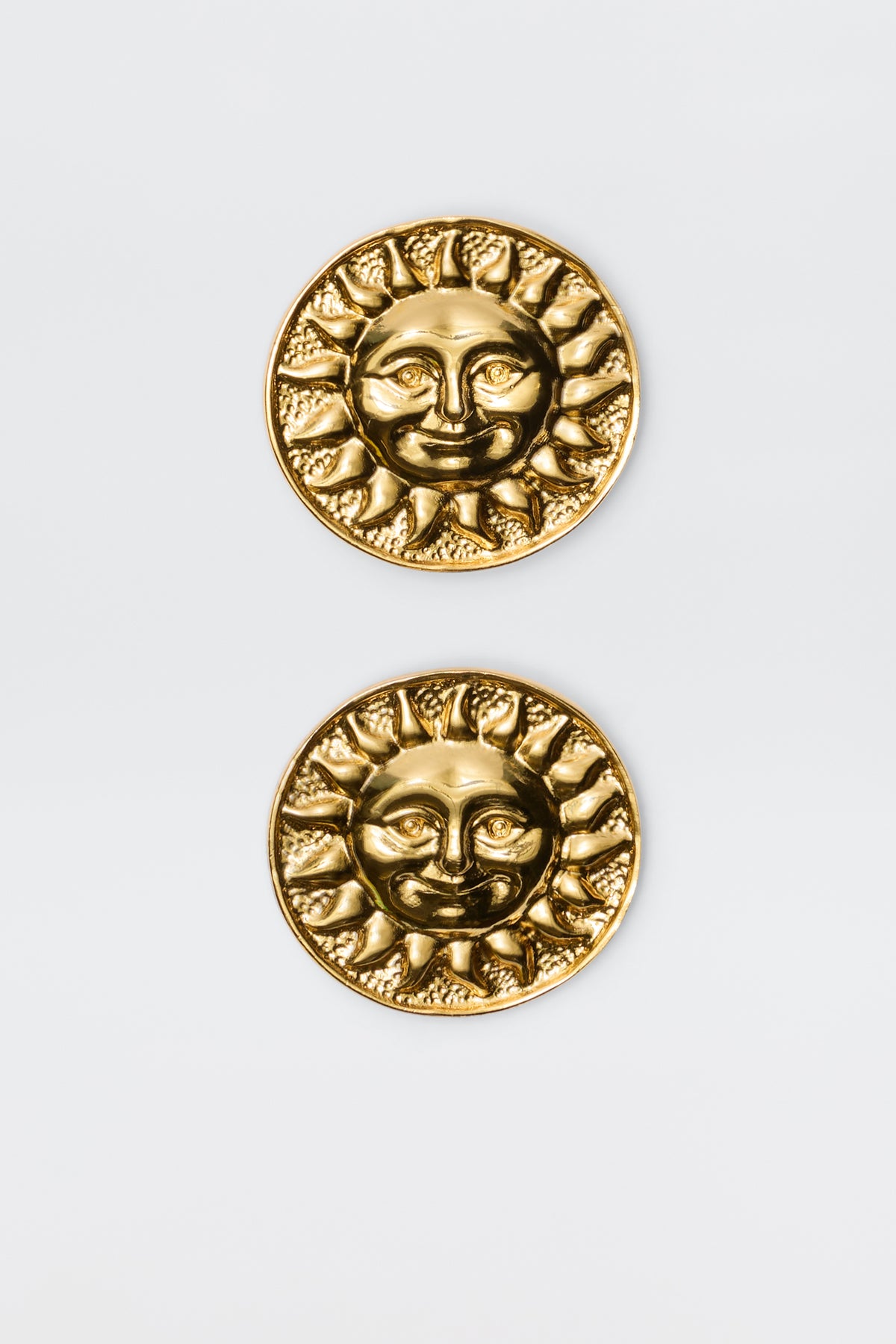 Introducing Noni - Gold: round gold-plated brass coins with embossed sun faces, rays, and intricate details. These vintage-inspired pieces are displayed vertically on a light gray background, resembling elegant round stud earrings.