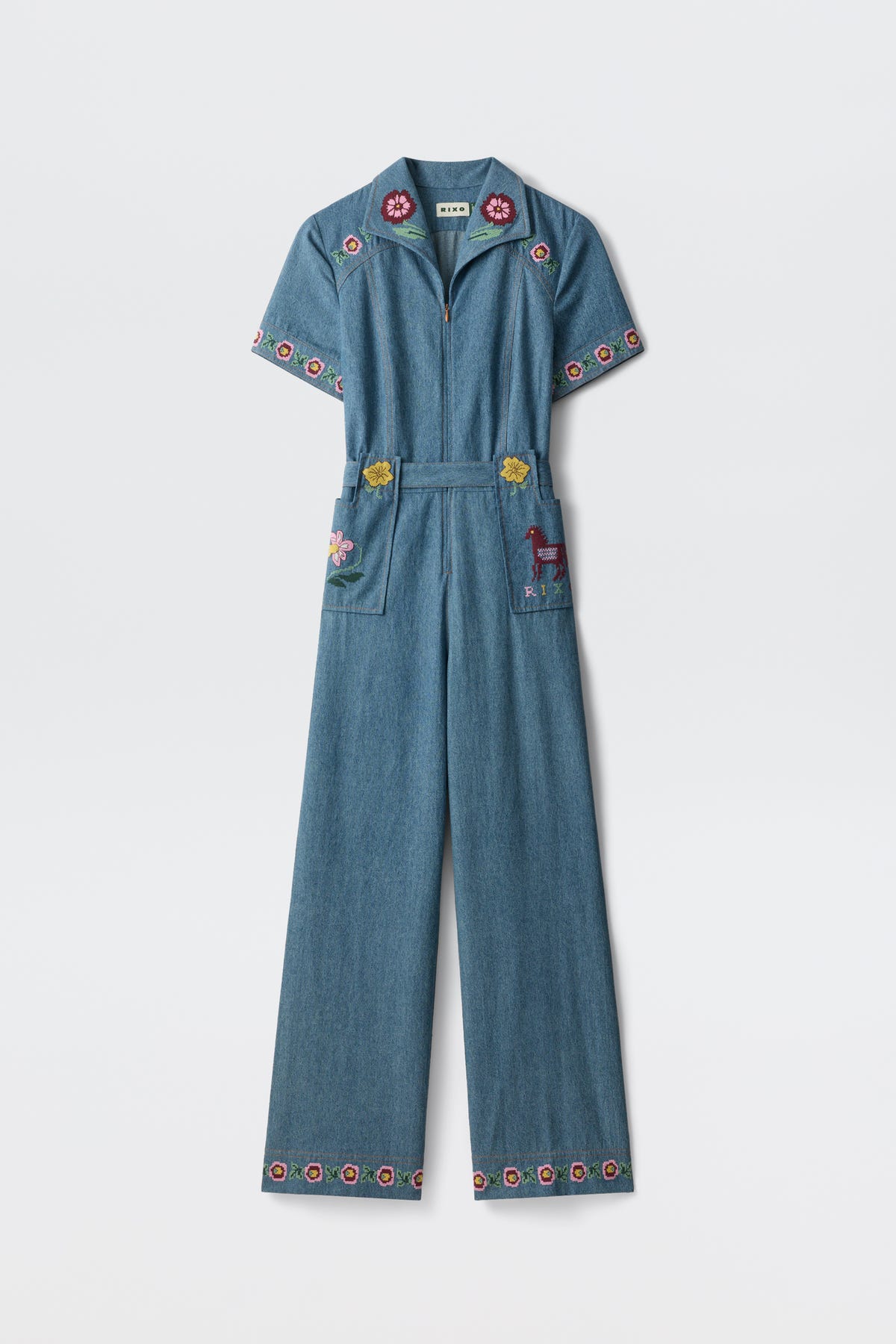 A blue denim jumpsuit, the Briana - Juliette Embroidery Blue, featuring short sleeves and wide legs with colorful floral embroidery on the collar, sleeves, pockets, and hem. One pocket is adorned with an embroidered deer. The jumpsuit is displayed against a plain white background.