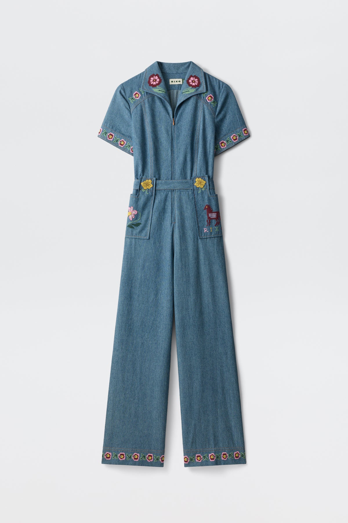 A blue denim jumpsuit, the Briana - Juliette Embroidery Blue, featuring short sleeves and wide legs with colorful floral embroidery on the collar, sleeves, pockets, and hem. One pocket is adorned with an embroidered deer. The jumpsuit is displayed against a plain white background.