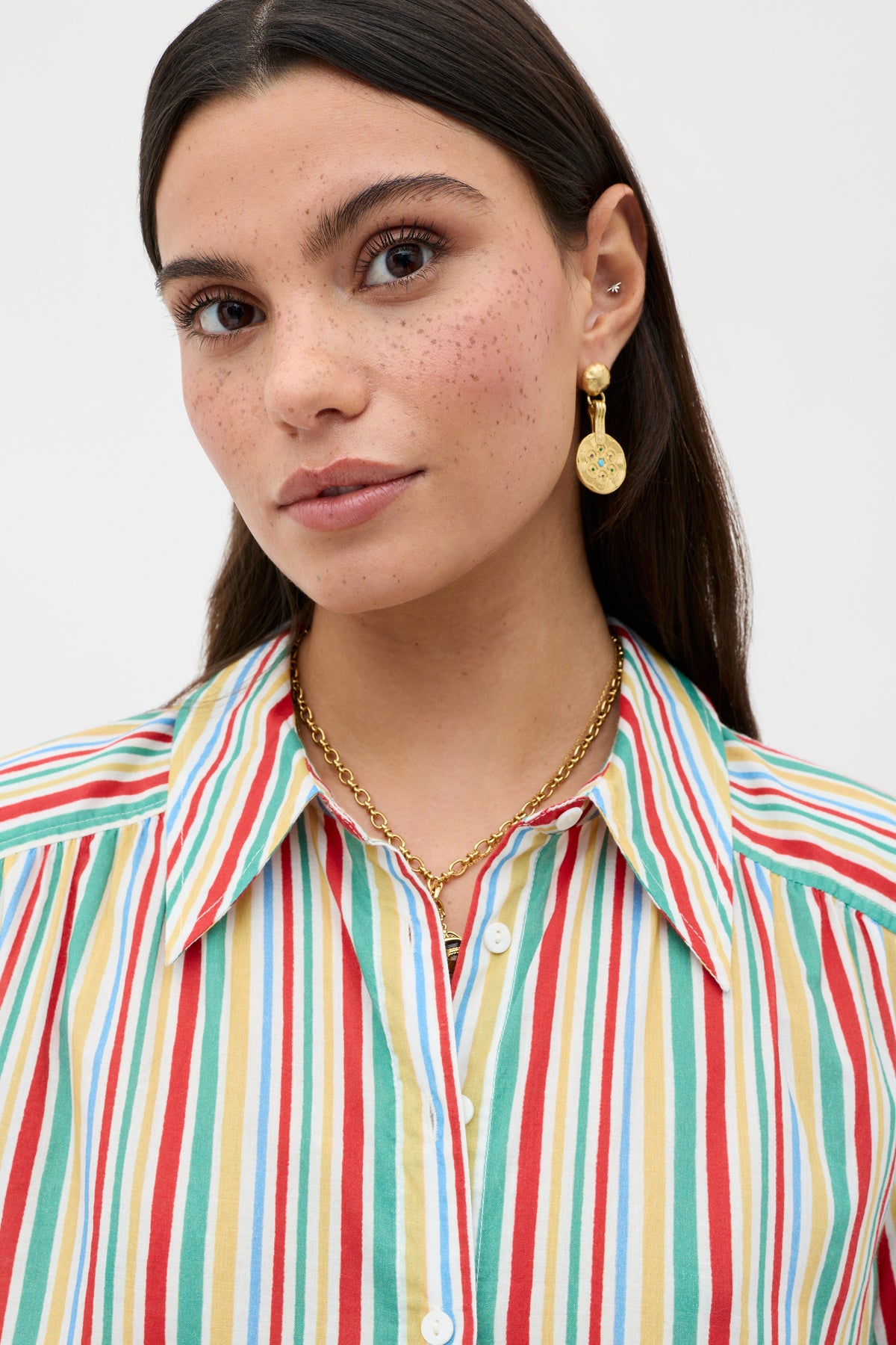 A woman with long dark hair and freckles wears a colorful striped shirt, gold chain necklace, and RIXO Anouk - Gold earrings, looking slightly to the side against a plain light background.