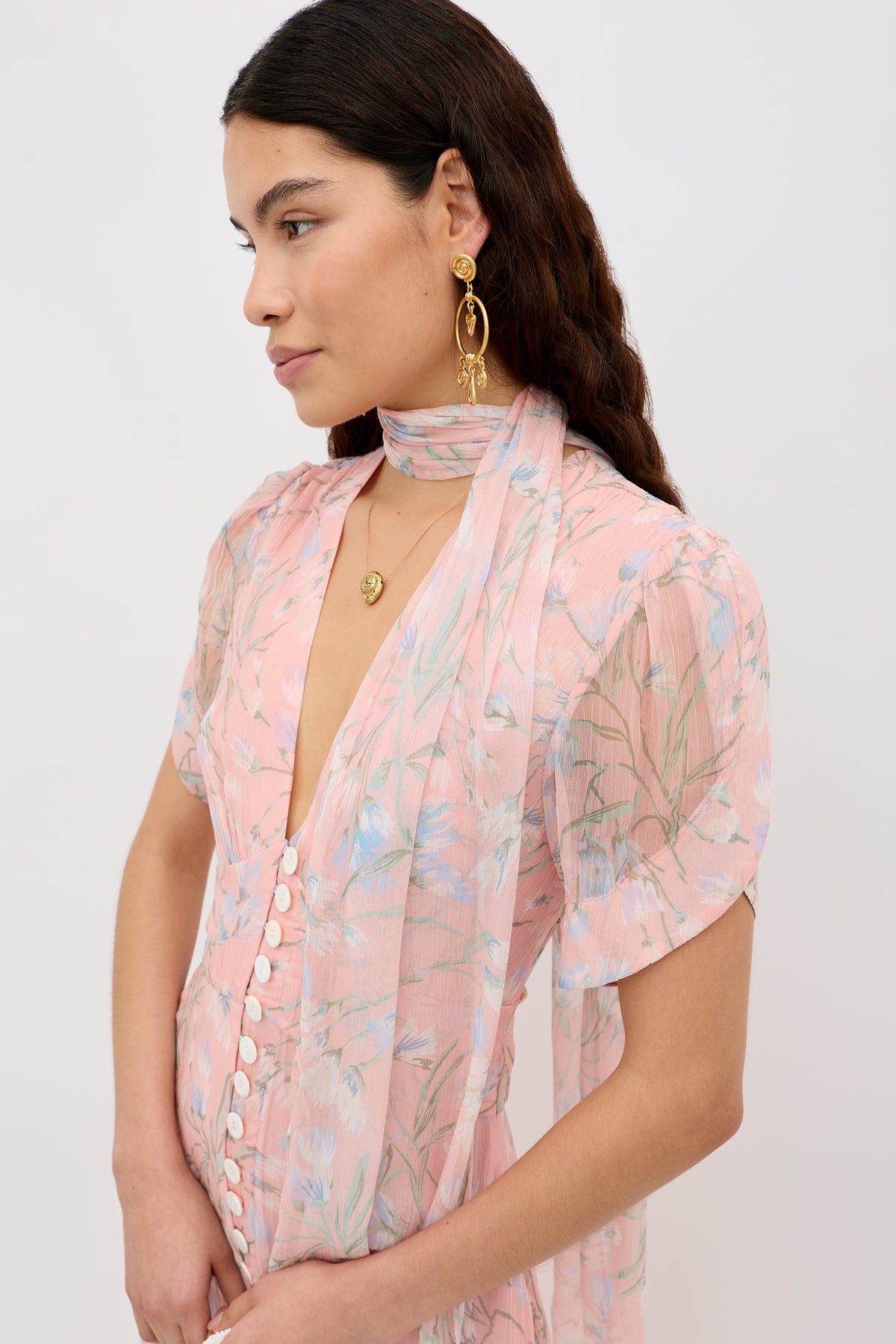 A woman with long, wavy brown hair wears the Julien - Watercolour Floral Pink dress with white buttons and a matching scarf. She accessorizes with gold drop earrings and a gold necklace, gazing off to the side against a plain background.