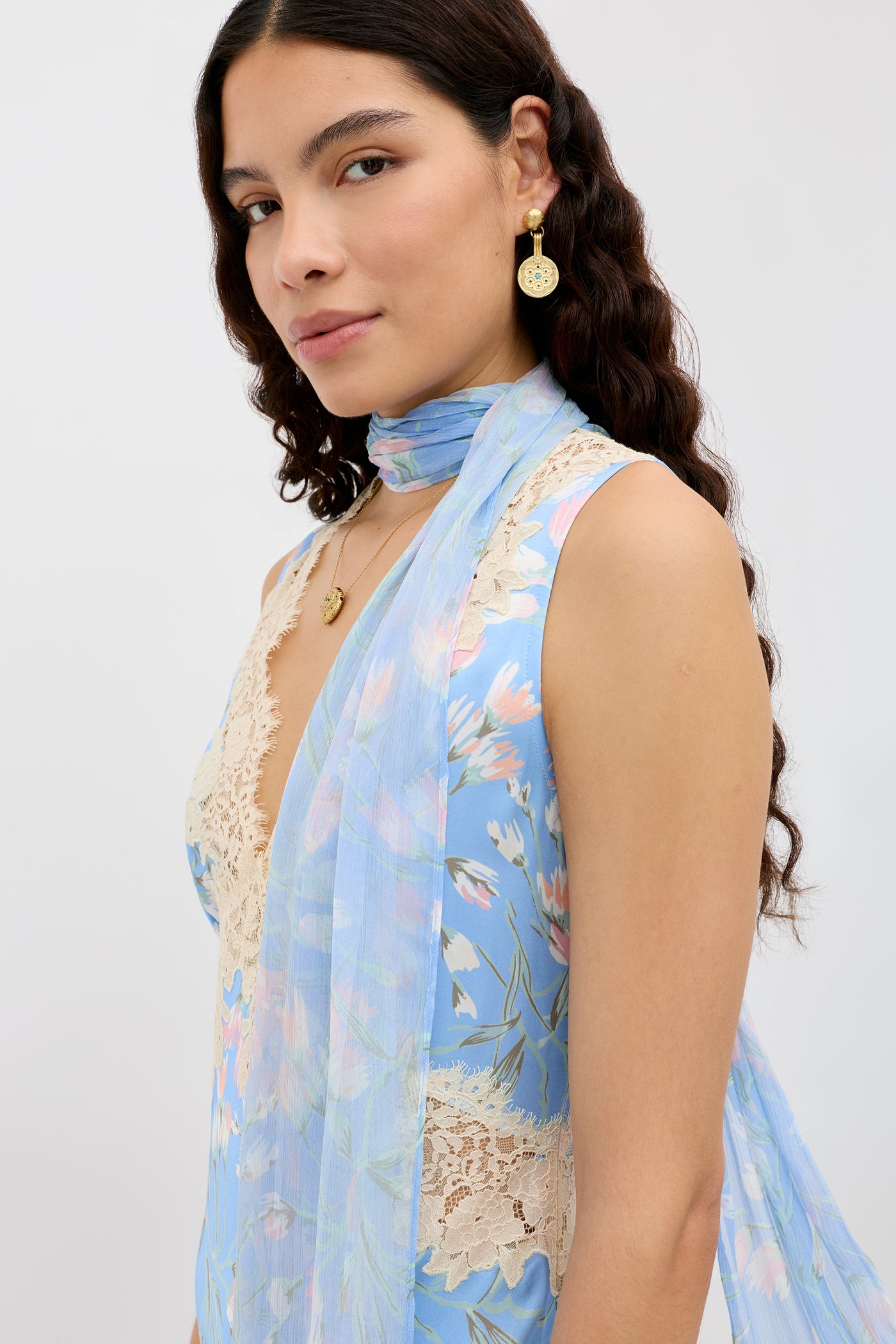 A woman with long wavy hair models the "Julien - Watercolour Floral Blue" sleeveless dress with lace accents and a sheer scarf, styled with gold earrings and necklace, posing before a plain light background.