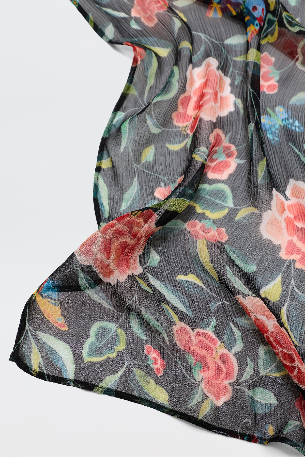 A close-up of Julien - Butterfly Floral Black shows sheer black fabric with large pink and red flowers, green leaves, and accents of blue and yellow, softly draped against white for a romantic scarf look.