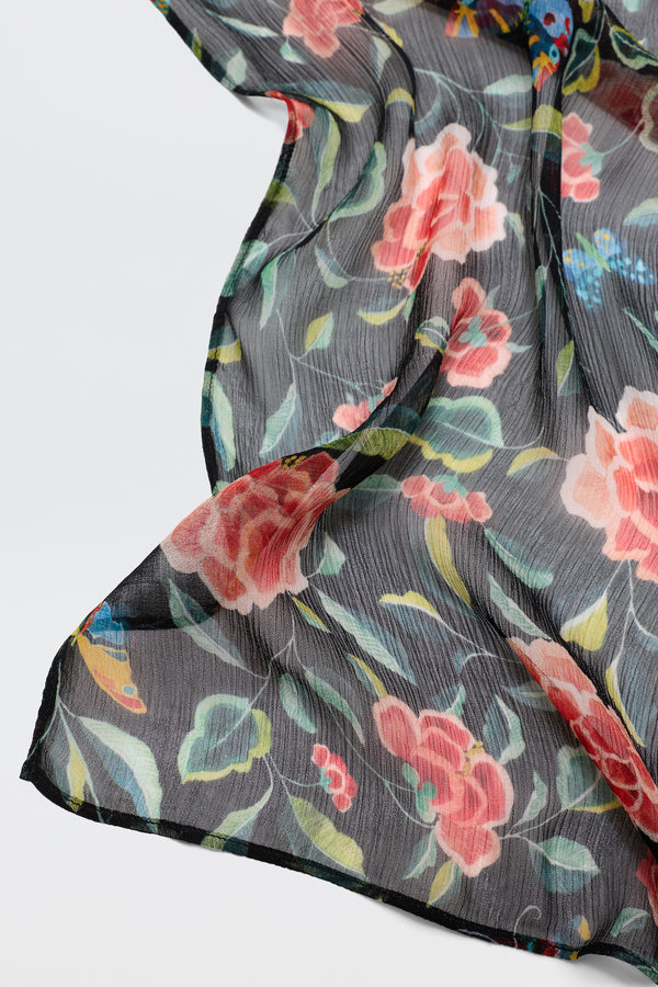 A close-up of Julien - Butterfly Floral Black shows sheer black fabric with large pink and red flowers, green leaves, and accents of blue and yellow, softly draped against white for a romantic scarf look.