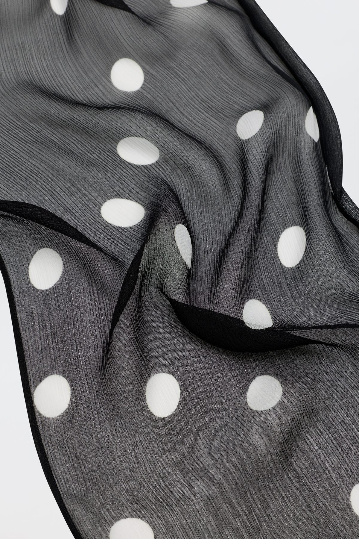Julien - Cosmos Spot Black is a sheer black fabric adorned with large white polka dots, slightly wrinkled and elegantly draped to create flowing, wavy folds on a light surface.