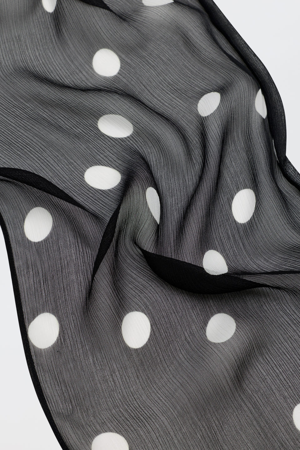 Julien - Cosmos Spot Black is a sheer black fabric adorned with large white polka dots, slightly wrinkled and elegantly draped to create flowing, wavy folds on a light surface.