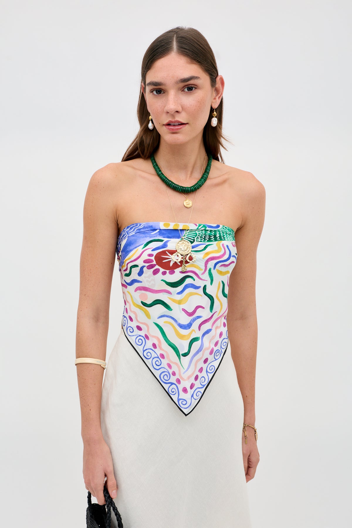 A woman with long brown hair wears the Menno - Mythos Iris Ivory, a colorful patterned strapless top styled like a scarf, with a cream skirt, layered necklaces, drop earrings, a bracelet, and holds a black bag against a plain light background.