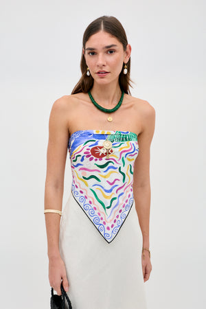A woman with long brown hair wears the Menno - Mythos Iris Ivory, a colorful patterned strapless top styled like a scarf, with a cream skirt, layered necklaces, drop earrings, a bracelet, and holds a black bag against a plain light background.