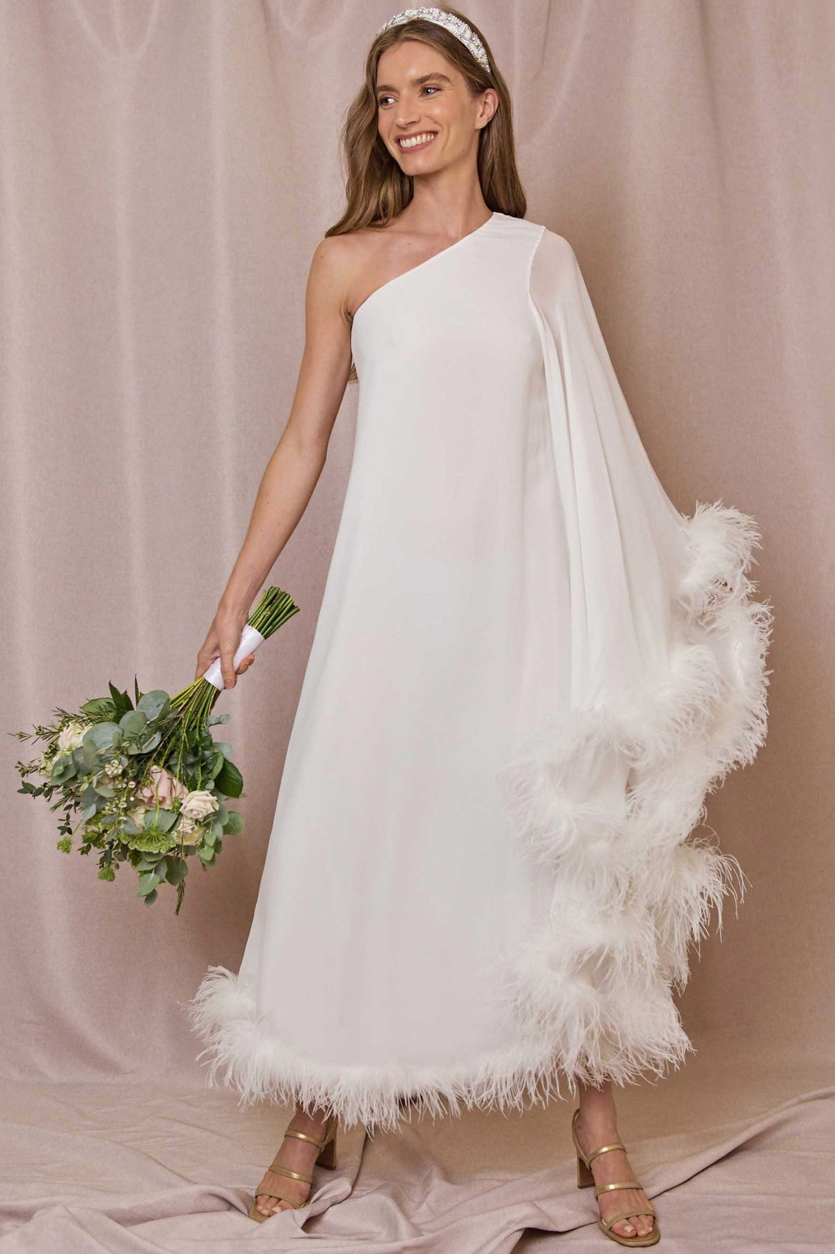 Wearing the Liza - Ivory Feathers dress with a one-shoulder neckline and feather trim, a woman in gold sandals and a headband smiles with a bouquet in hand before a beige backdrop.