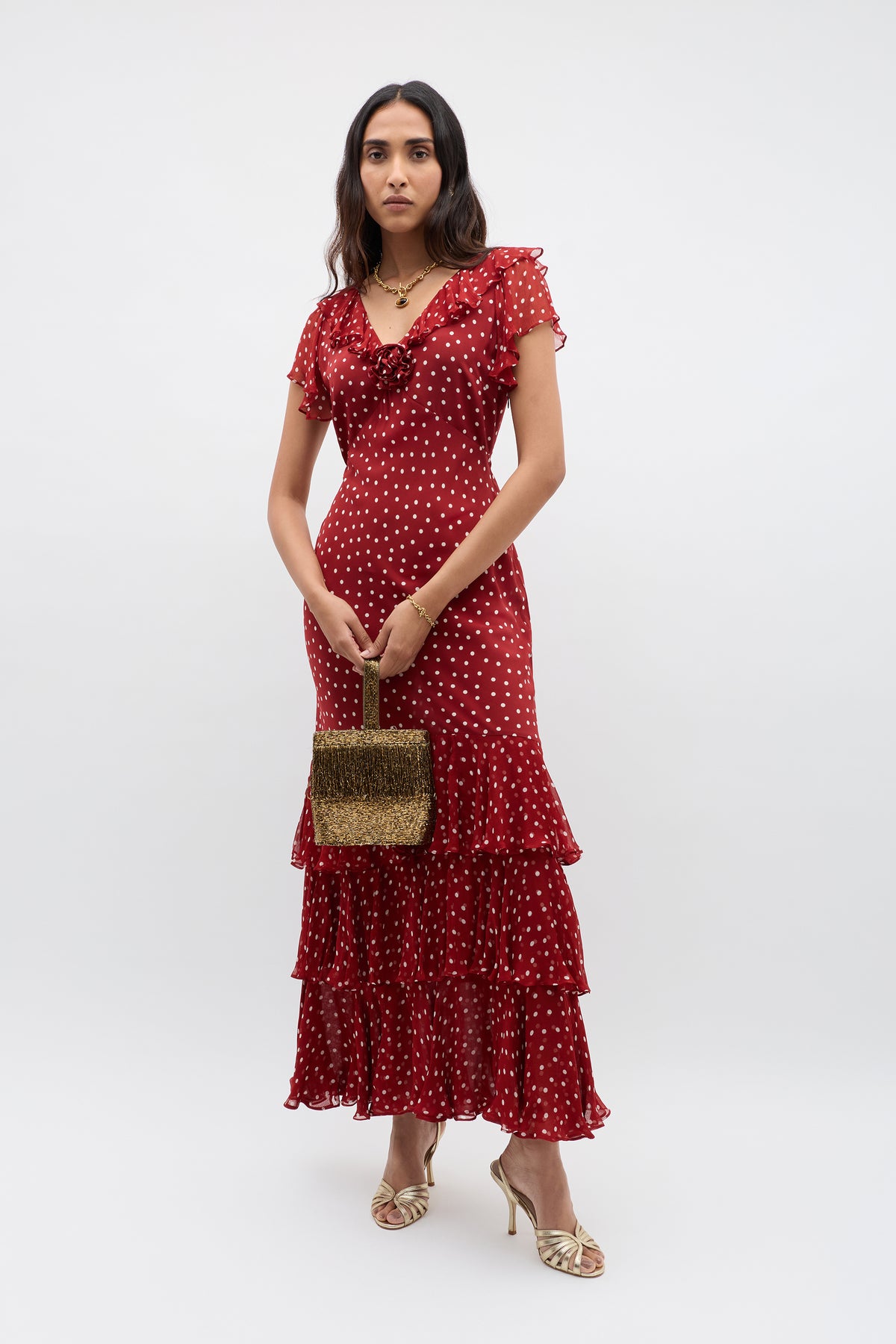 A woman in a red, short-sleeved, ruffled maxi dress with white polka dots holds a silk-lined occasion bag with a rounded top handle and wears Zenni - Beaded Gold strappy heels, standing against a plain white background.