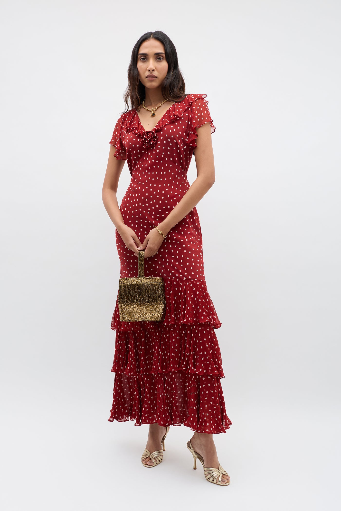A woman in a red, short-sleeved, ruffled maxi dress with white polka dots holds a silk-lined occasion bag with a rounded top handle and wears Zenni - Beaded Gold strappy heels, standing against a plain white background.