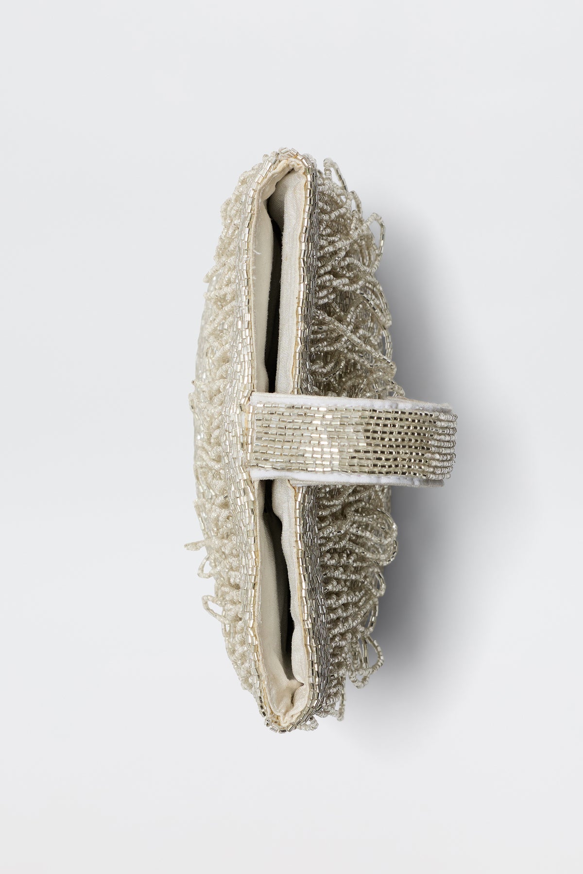 The Zenni - Beaded Silver is an off-white occasion bag with intricate beadwork, a decorative center strap, and a rounded top handle, shown from the side against a plain white background.