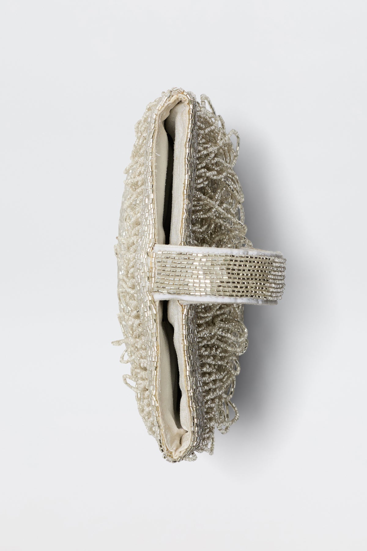 The Zenni - Beaded Silver clutch purse is shown from above with a textured, fringed exterior and a strap decorated with shiny beads. The occasion bag is open, revealing its light-colored interior.