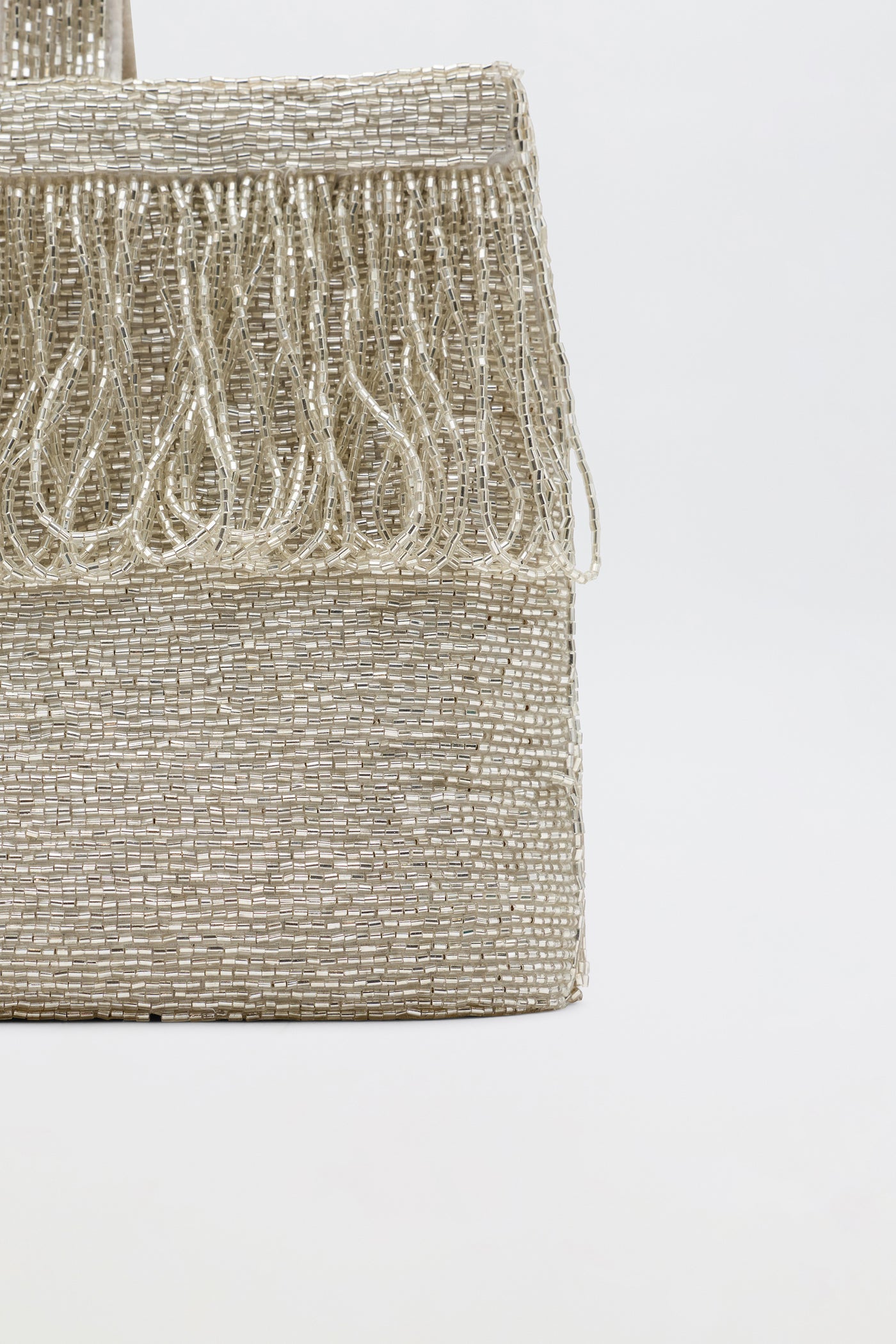 Close-up of Zenni - Beaded Silver rectangular metallic clutch with fringe along the top edge and a rounded top handle, set against a plain light background.