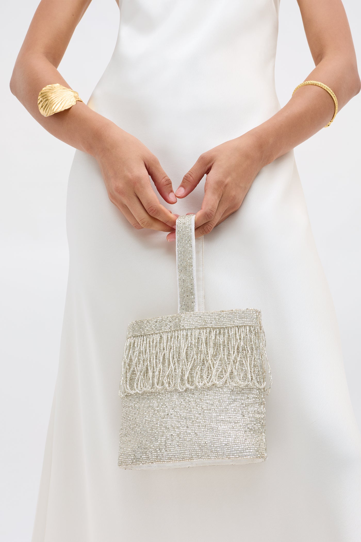Wearing a white satin dress, a person holds the Zenni - Beaded Silver bag with fringe and a rounded top handle, styled with a gold shell bracelet and simple gold bangle, set against a plain light background.