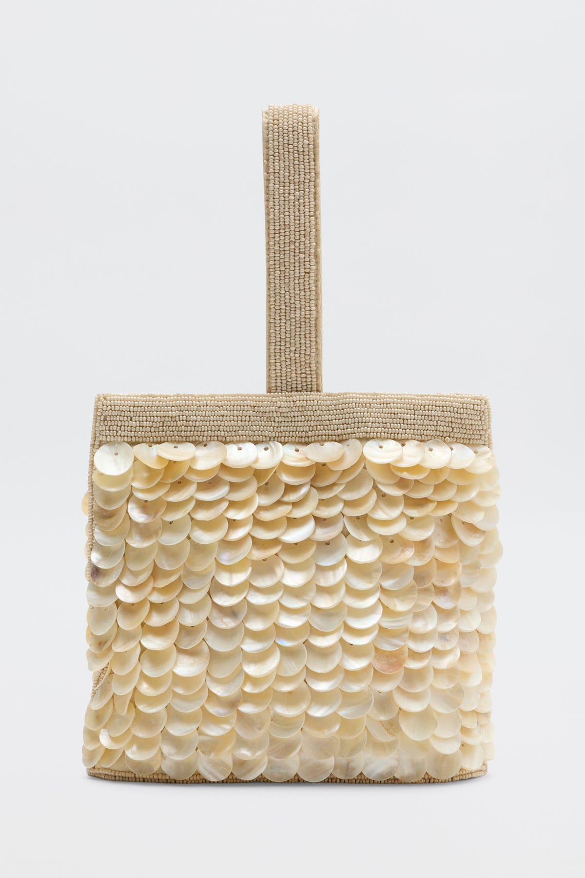The Zenni - Shell is a small, square beige handbag featuring layered shiny shell discs on the front and a wide woven handle, shown on a plain white background.