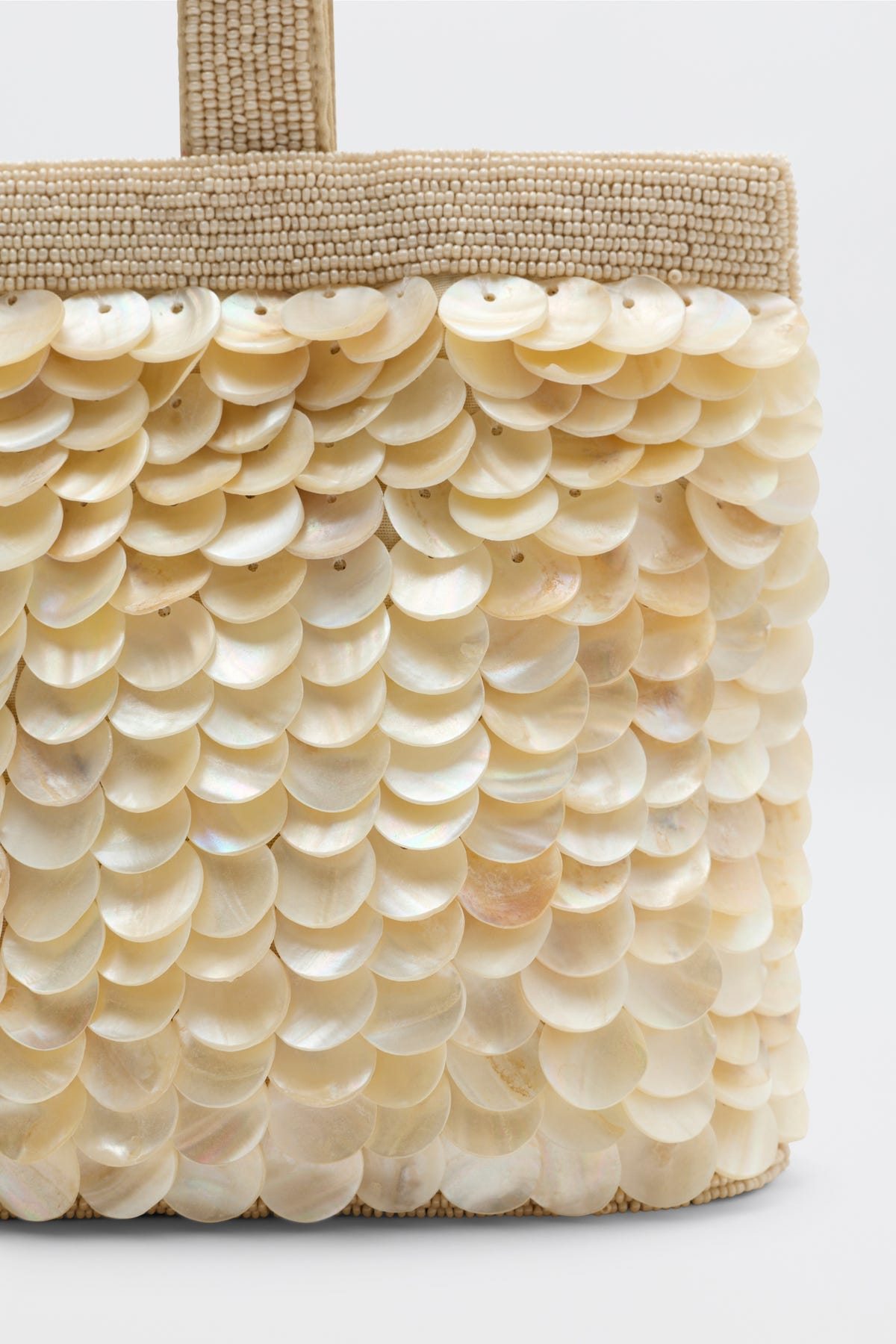 A close-up of the Zenni - Shell bag, crafted in beige weave and adorned with overlapping, shiny mother-of-pearl discs that form a textured, shimmering surface.