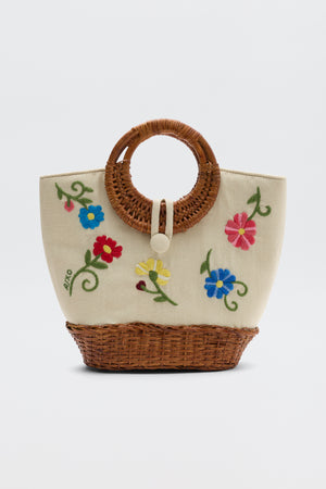 The Zelene - Ombre Floral Embroidery Cream is a beige hand-crafted bag with circular wicker handles, colorful floral embroidery with green stems, and a front button closure.