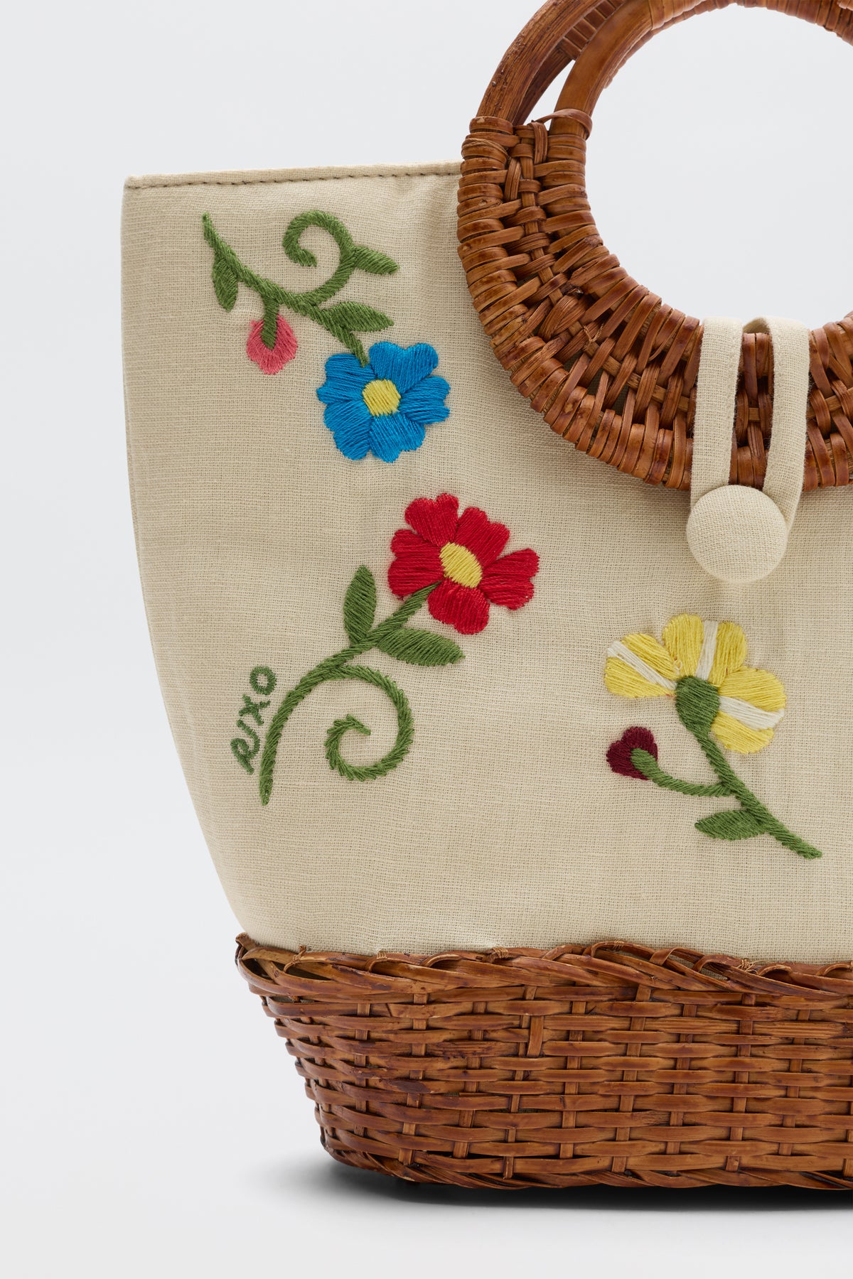 The Zelene - Ombre Floral Embroidery Cream is a hand-crafted beige fabric bag with a wicker handle, showcasing colorful red, blue, and yellow floral embroidery with green stems and leaves.