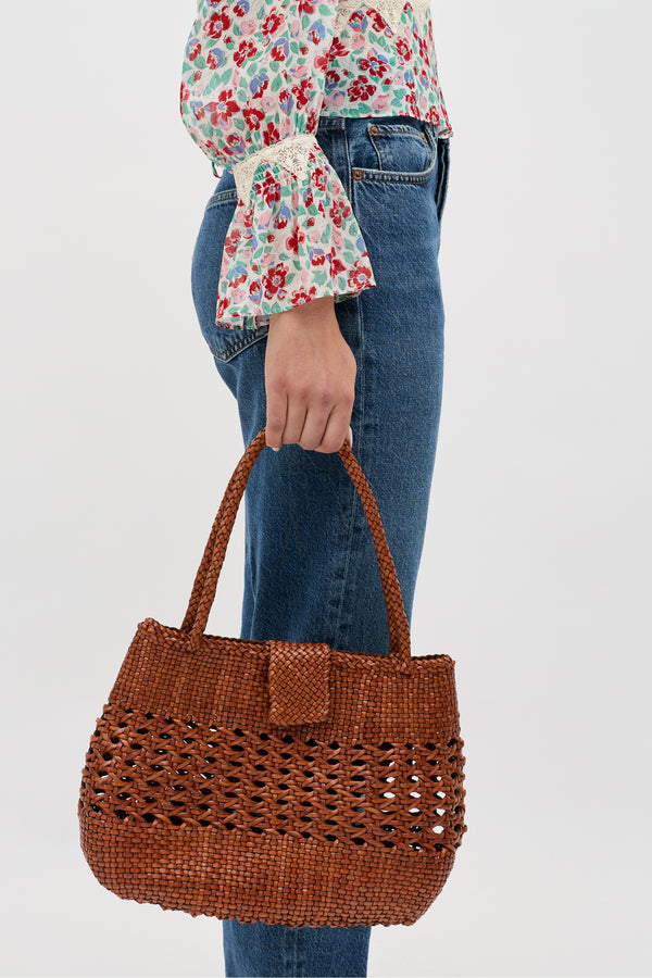 A person in blue jeans and a floral blouse holds the Niamh - Tan bag, featuring two handles and a textured open-weave design, highlighting its vintage woven charm against a plain background.