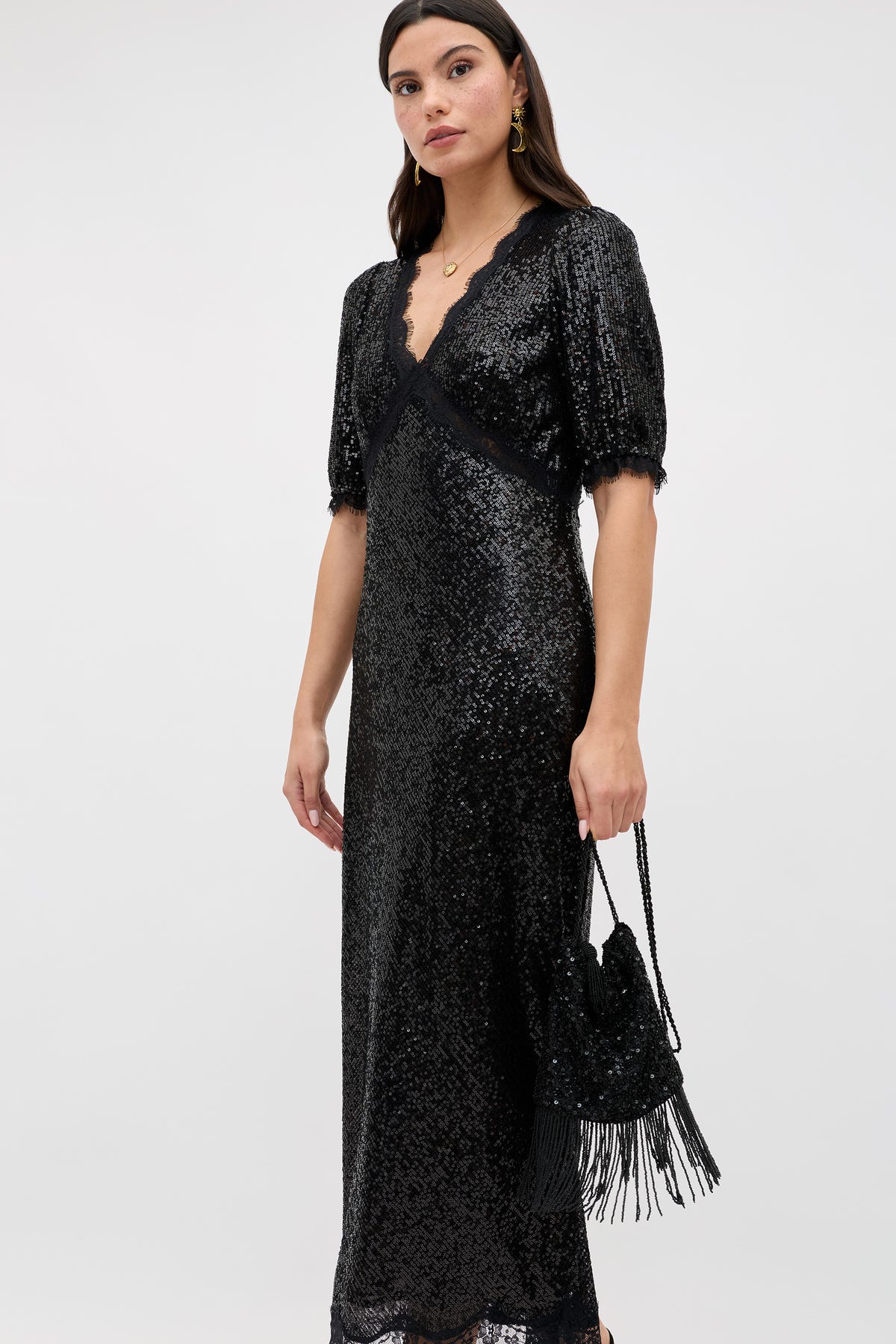 A woman wears the Orla - Vintage Scatter Embellishment Black dress, featuring black sequins and short sleeves. She holds a matching black fringed bag and stands slightly angled against a plain light background.