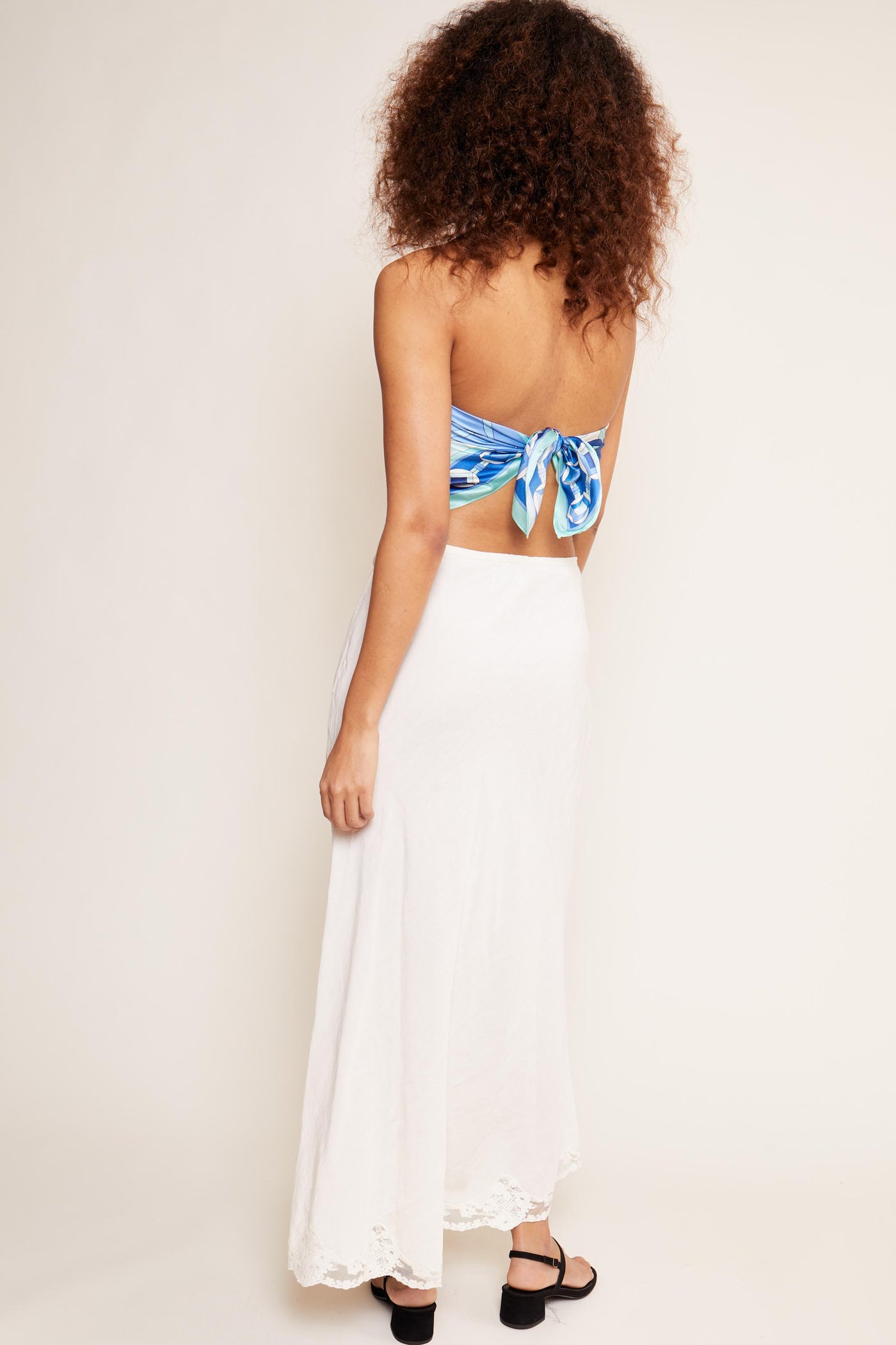 A woman with curly hair faces away, wearing the Crystal - Cream linen skirt with lace trim, a blue strapless top tied at the back, and black sandals against a neutral background.
