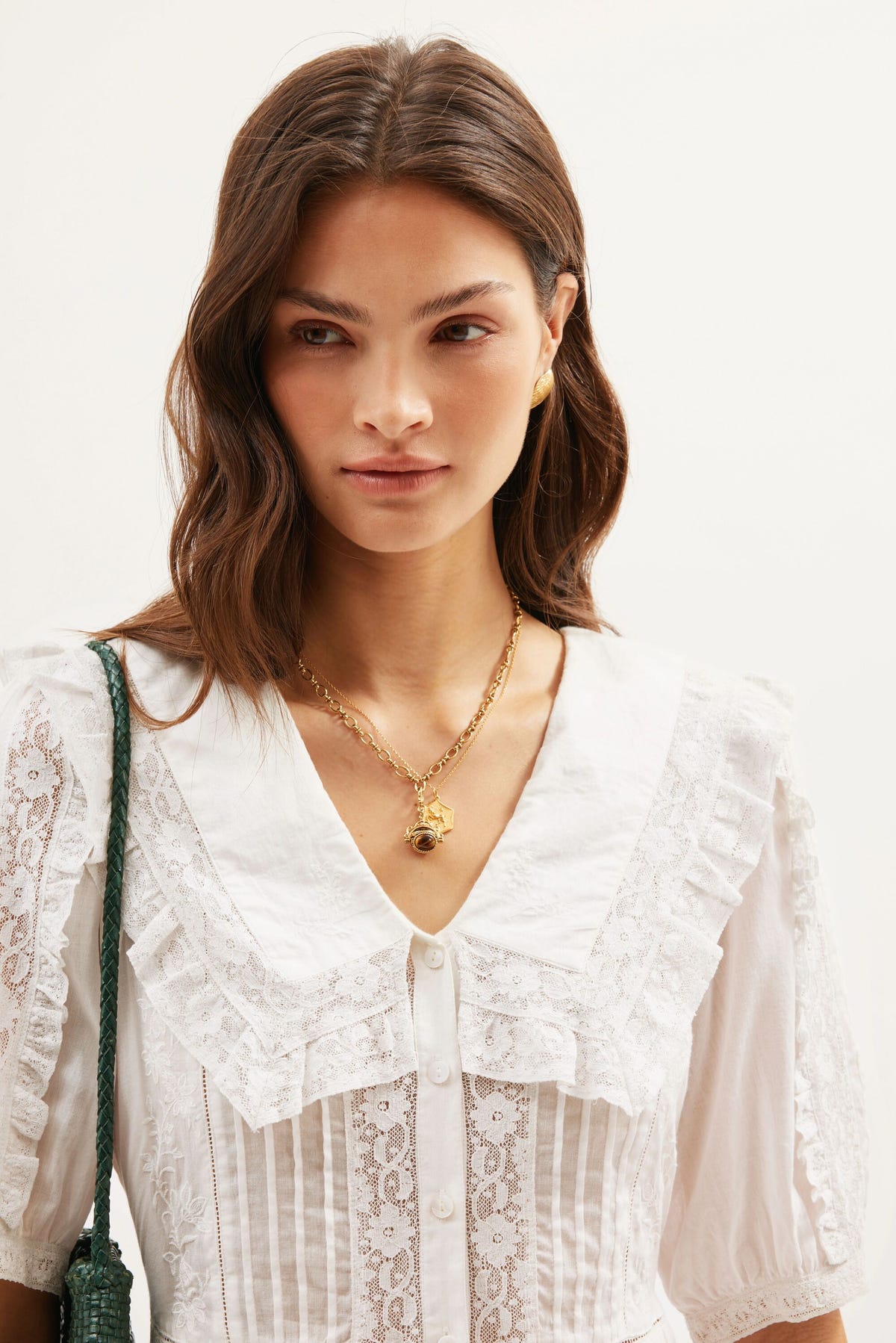 A woman with wavy brown hair wears the Dove - Pansy Embroidery White blouse, featuring lace trim and cotton-voile fabric. She accessorizes with a gold necklace, gold earrings, and carries a green shoulder bag. Her expression is neutral, set against a plain, light-colored background.