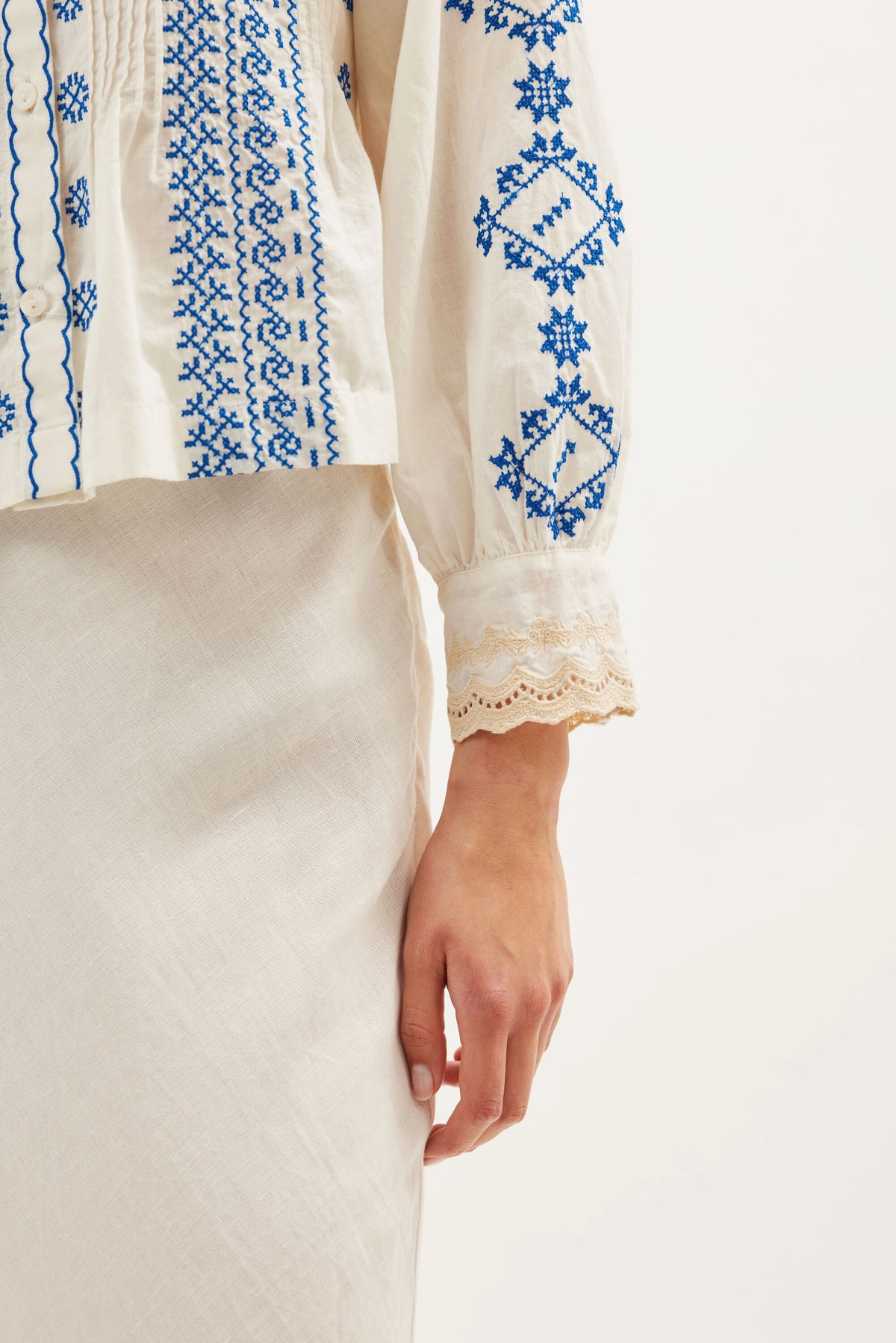 A close-up shows a person in the Percy - Mila Embroidery Cream: an off-white skirt and vintage-style blouse with blue embroidery and cotton lace sleeve trim. Only the lower half of the feminine blouse and one arm are visible.