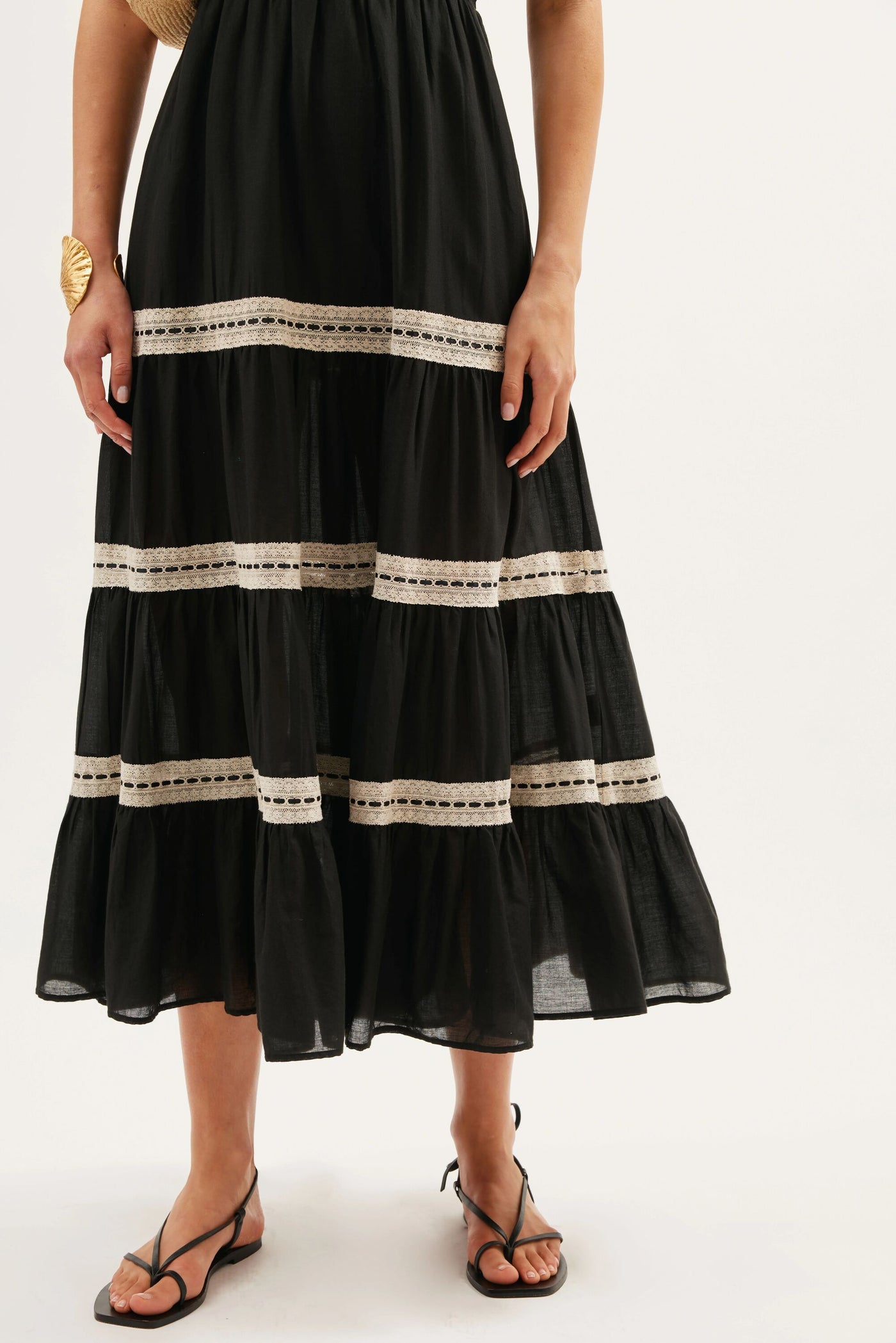 A person wearing the Aubrie - Black ankle-length skirt with beige crochet lace trim, brown sandals, and a gold leaf bracelet, standing against a plain white background.