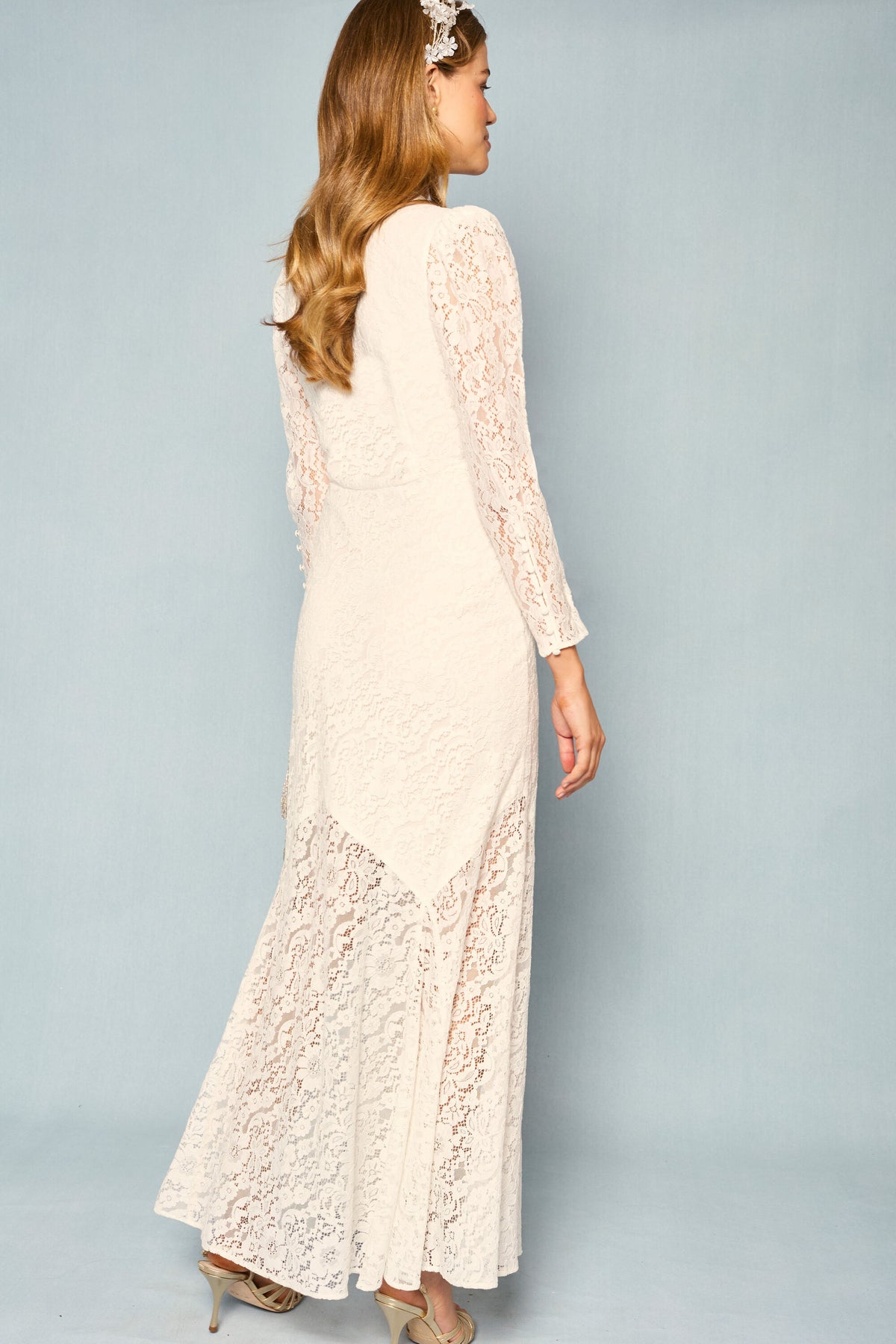 A woman with long, wavy hair wears the Fleur - Ivory: a vintage-inspired, floor-length corded lace dress with a fit-and-flare silhouette and beige heels. She stands against a light blue background, facing away and looking to the side.